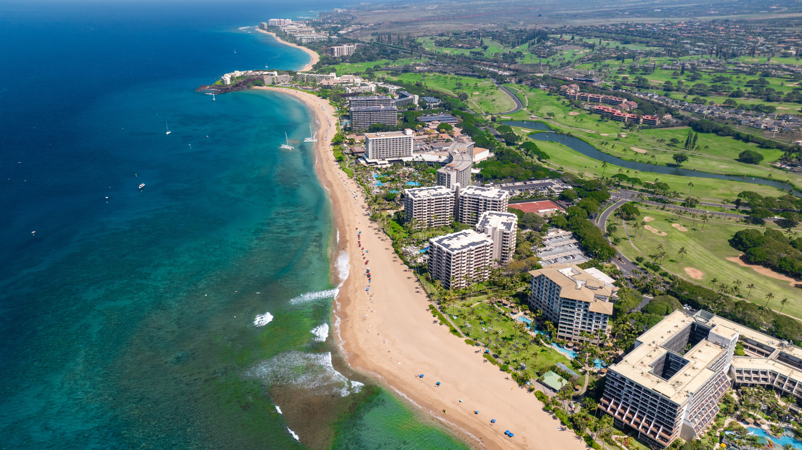 Aerial view of a beachfront resort with high-rise hotels lining a sandy coastline and a green inland golf course area.