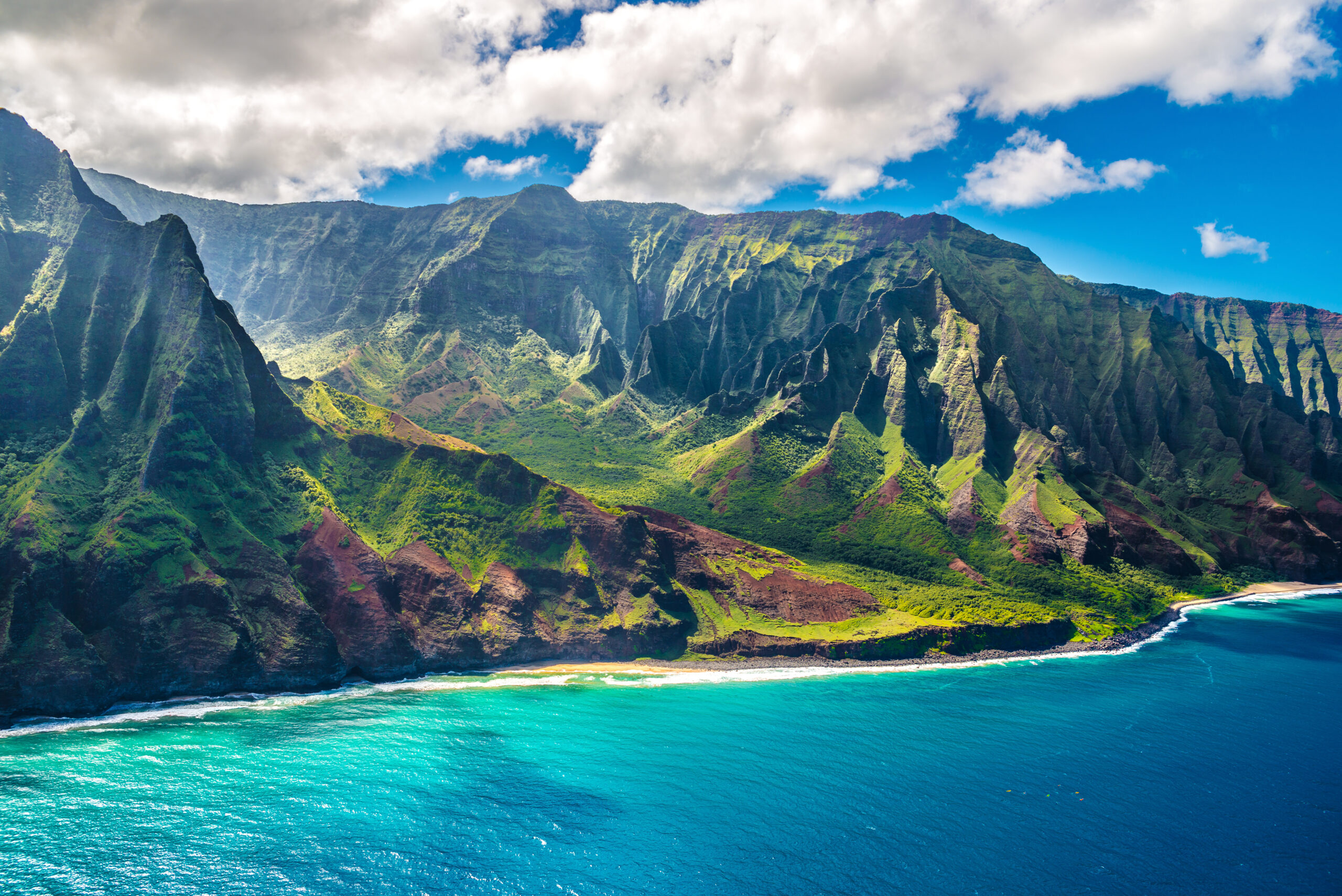 Steep green cliffs rise from a turquoise ocean under a blue sky with fluffy clouds along a rugged coastline.