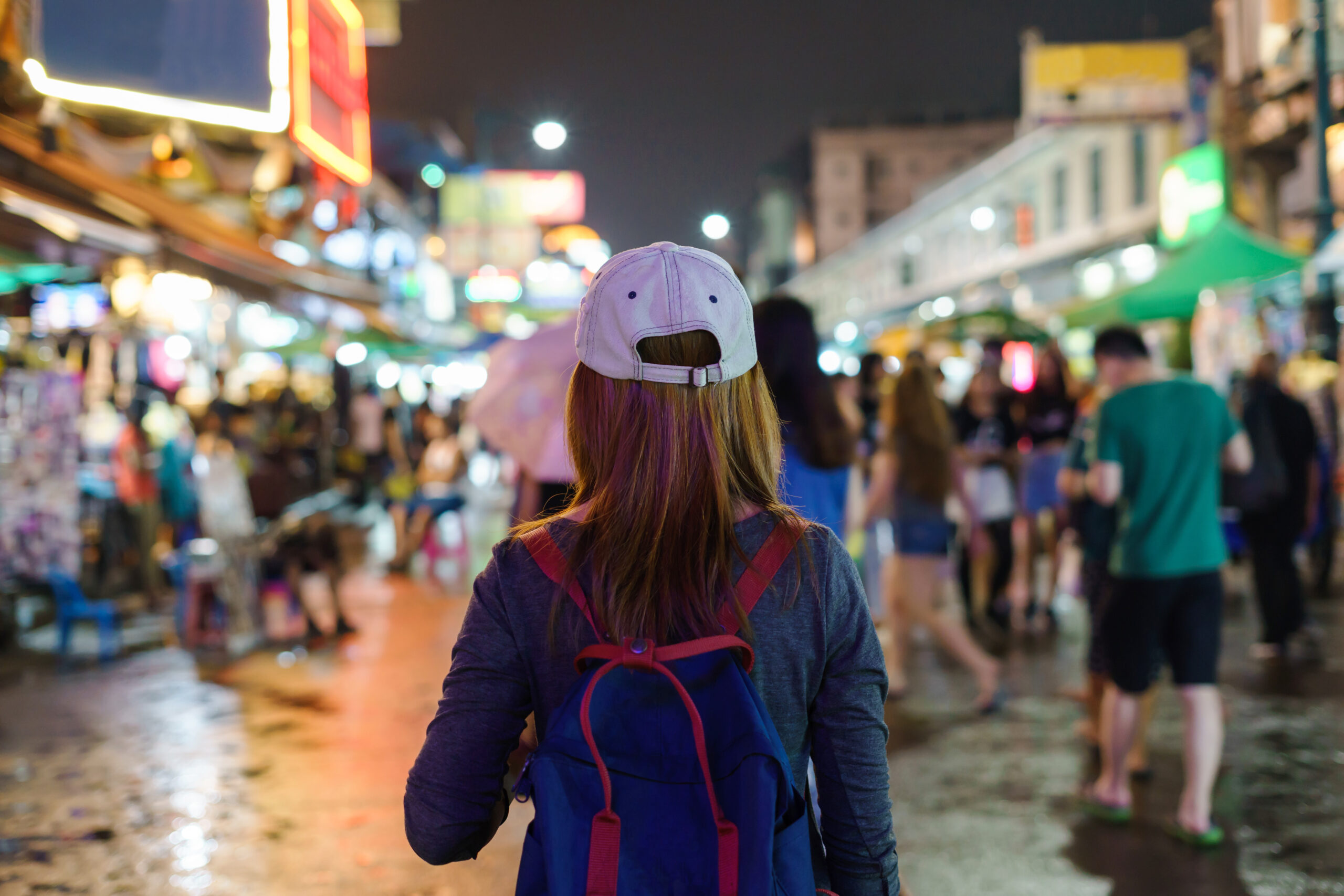 Person seen from behind wearing a light gray cap and blue backpack in a busy nighttime market with colorful neon lights and blurred crowd.