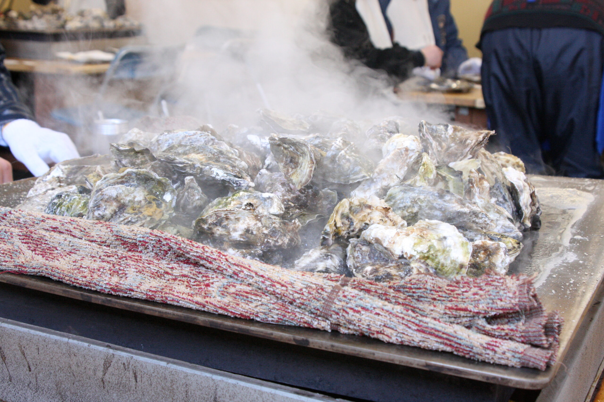 Pile of steaming oysters or shells on a grill, with a gloved hand nearby and a woven mat in the foreground