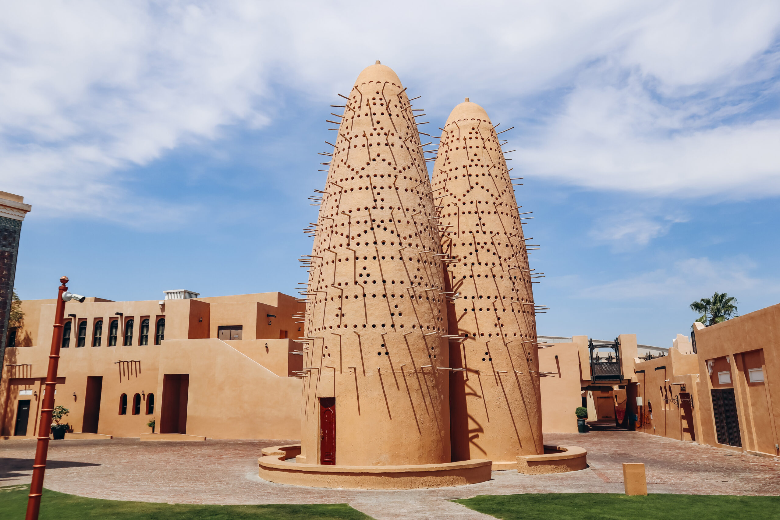 Two tall beige cone towers with perforations and wooden spikes in a sunlit desert courtyard against a blue sky.