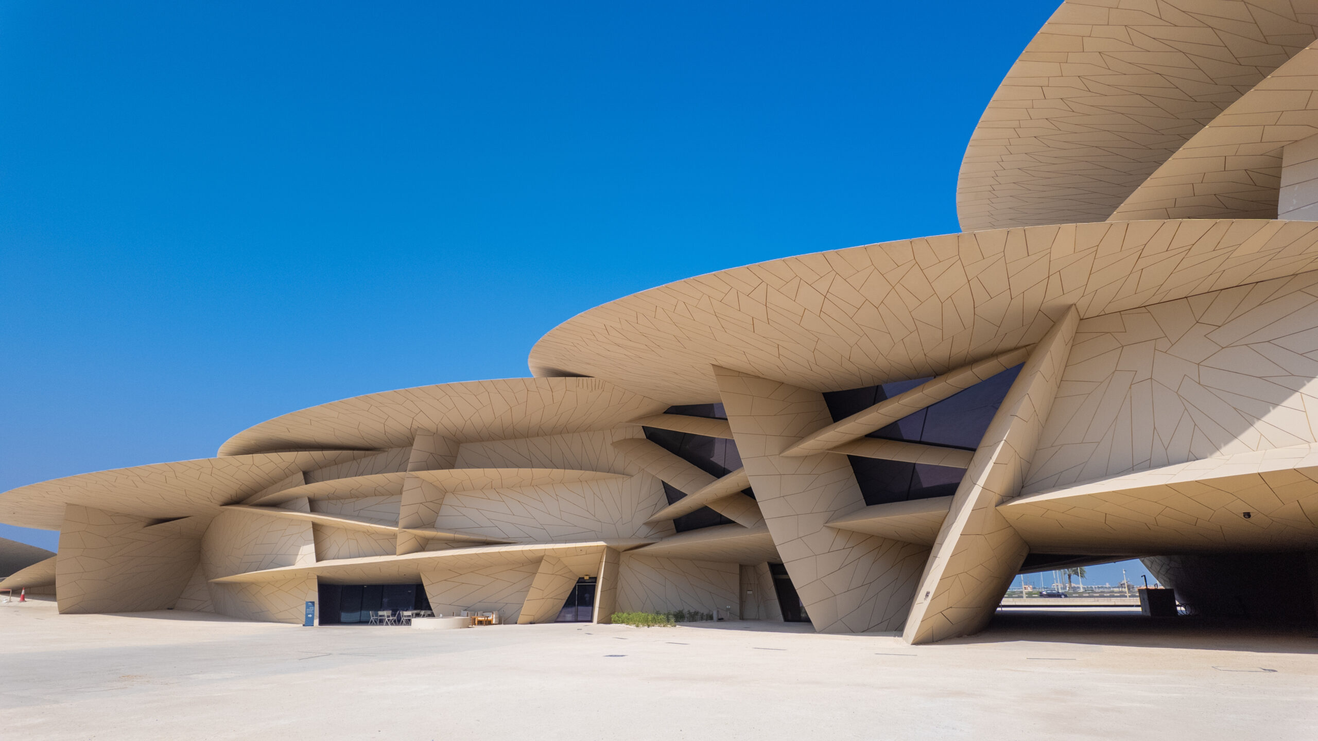 Beige futuristic building with curved layered platforms under a clear blue sky, showing geometric tiled surfaces and arches.