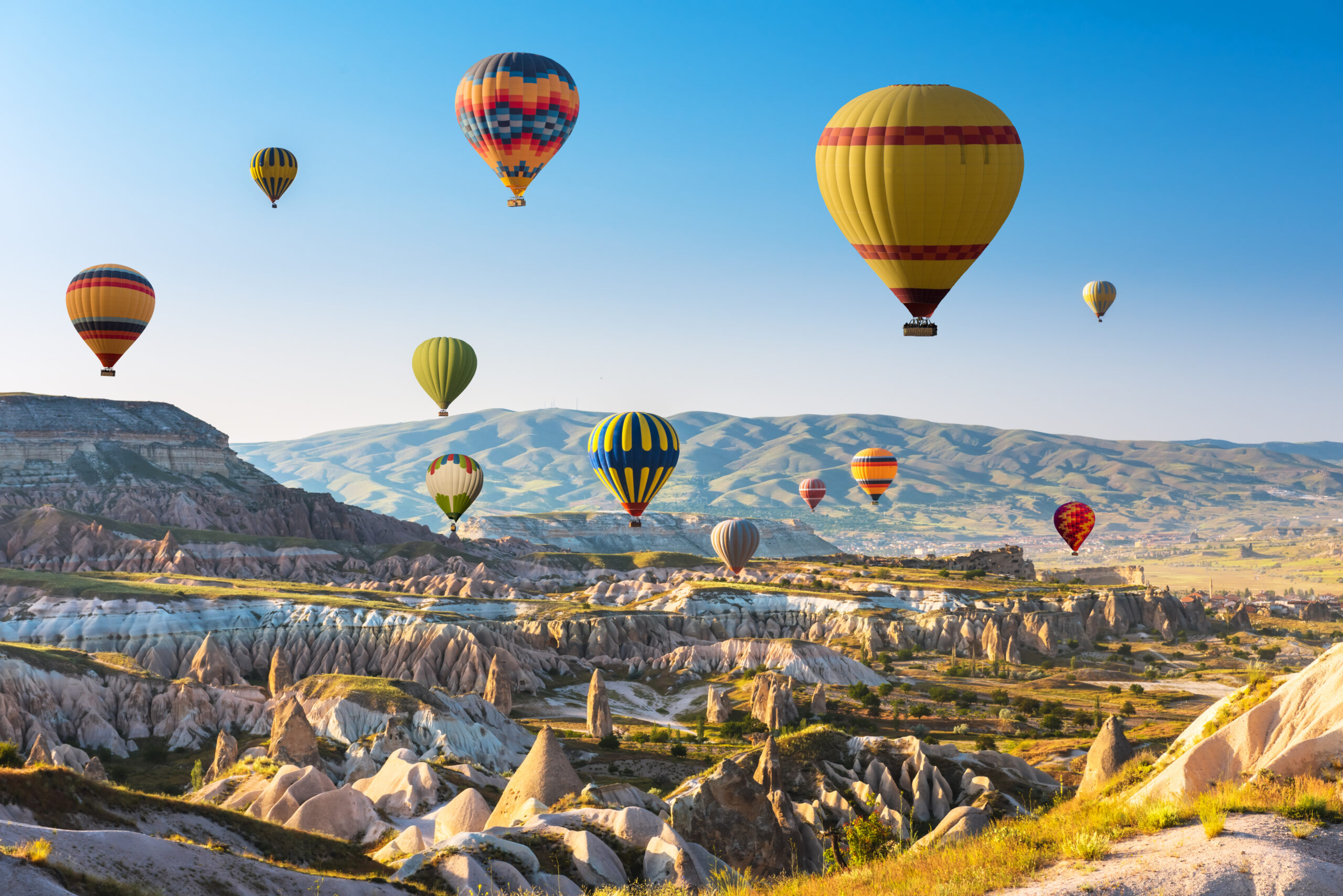 Colorful hot air balloons floating over a surreal rocky valley under a clear blue sky at dawn/dusk.
