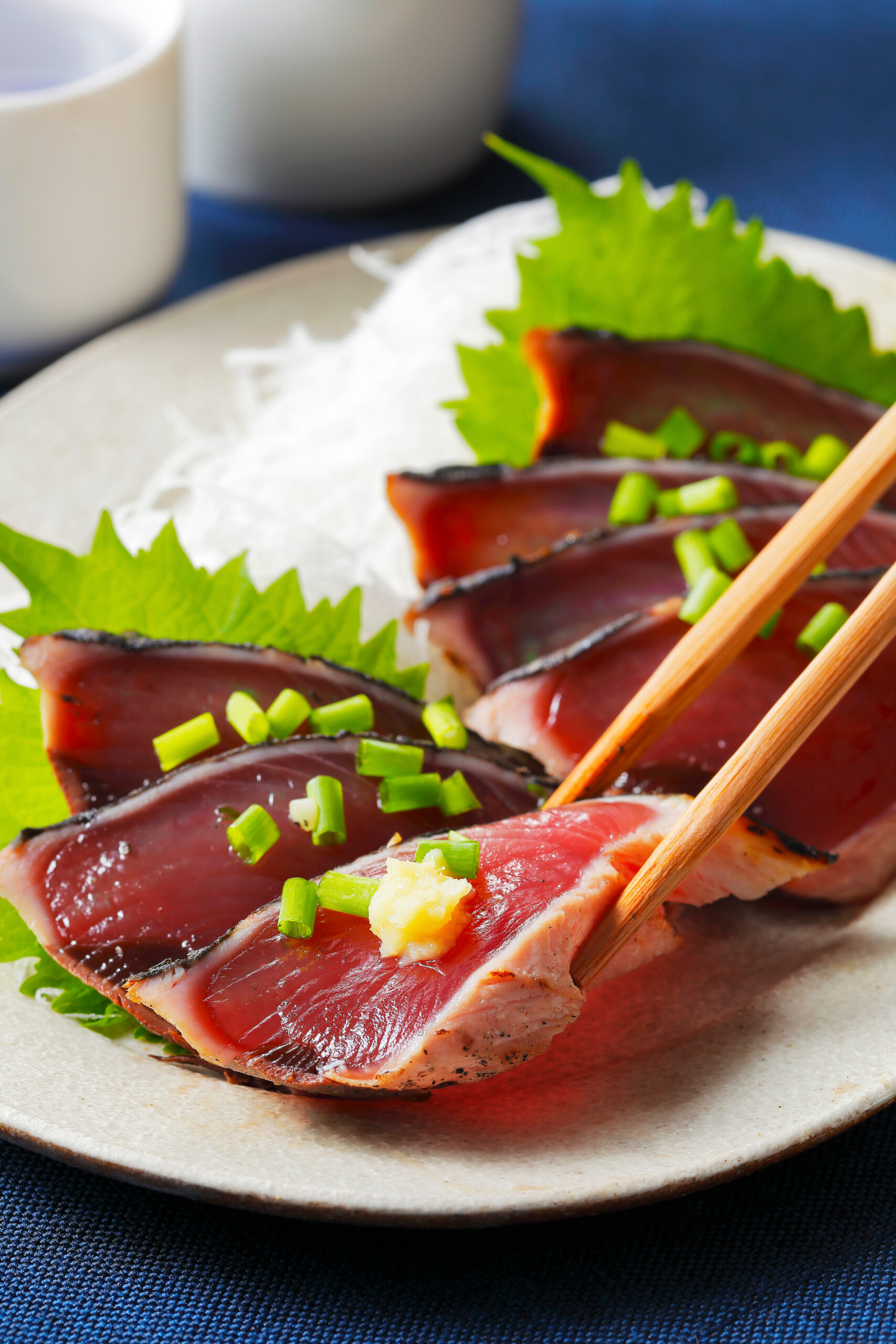 Sliced seared tuna with green onion garnish on a white plate, with fresh greens and chopsticks nearby.