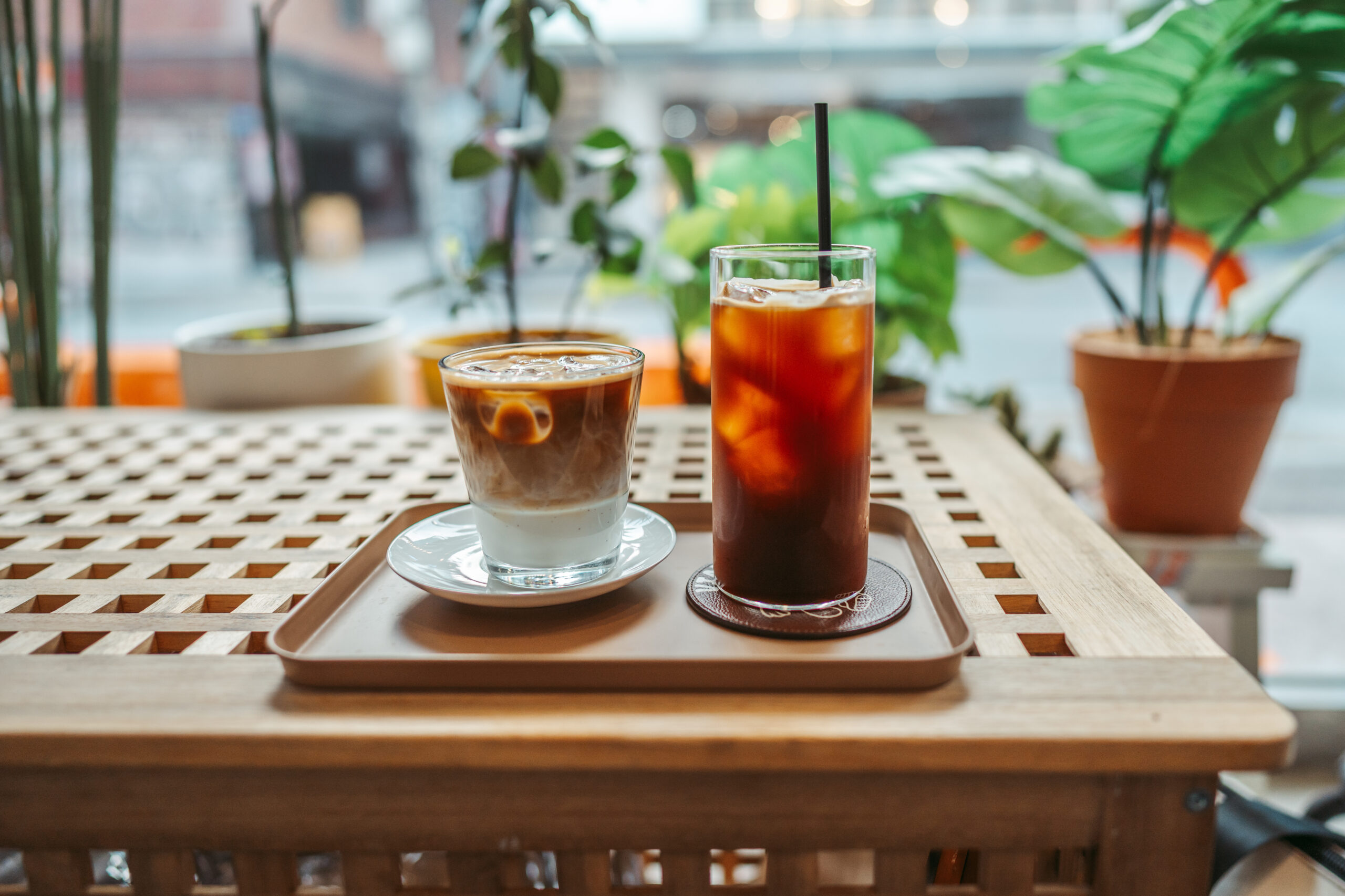 Two iced drinks on a tray at a cafe: a layered iced latte and a tall glass of iced tea with a straw, with greenery in the background.