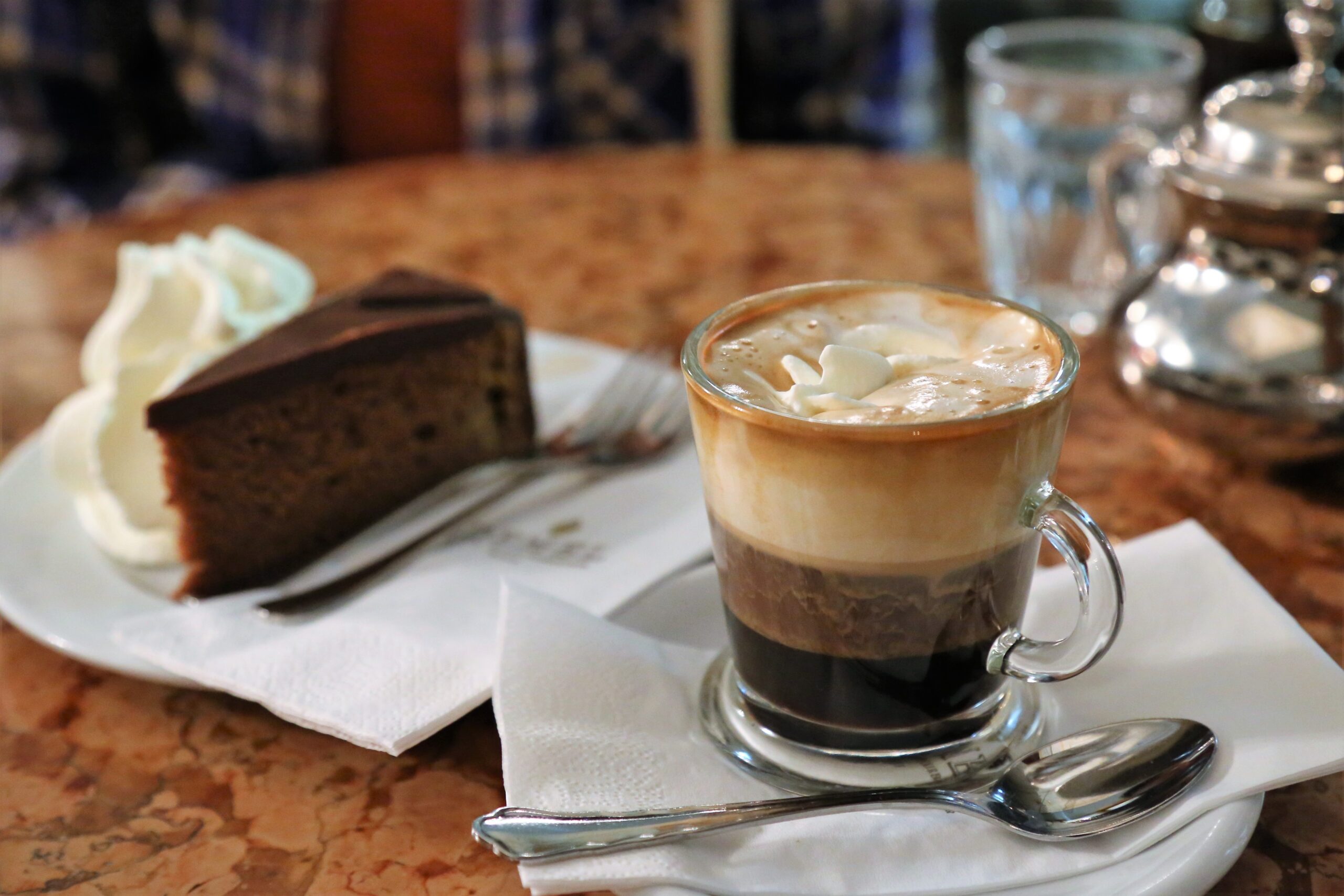 Cup of layered coffee with whipped cream on top and a slice of chocolate cake on a plate beside it on a marble table, napkin and utensils included.