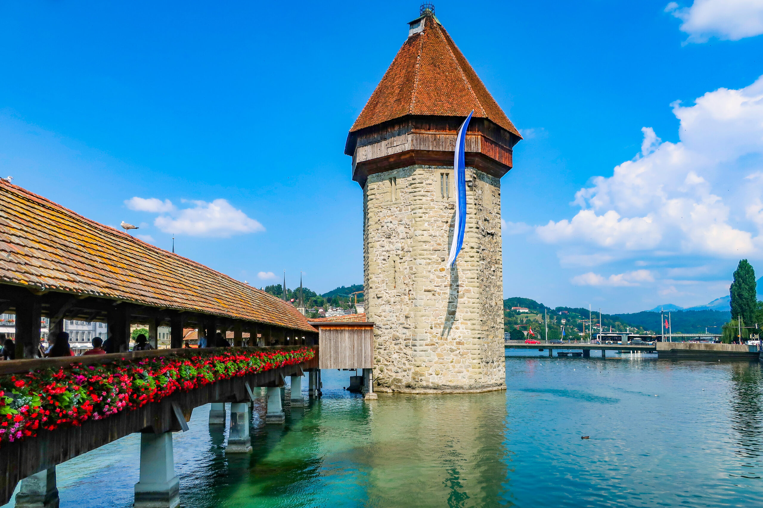 Stone lakeside tower with a red-tiled, conical roof beside a covered wooden walkway lined with red flowers.