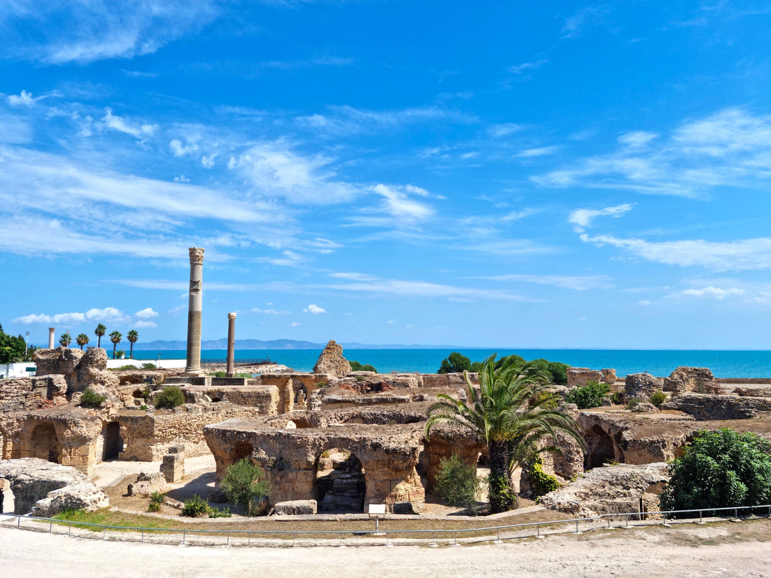 Ancient stone ruins with tall columns along a coastal landscape under a bright blue sky.