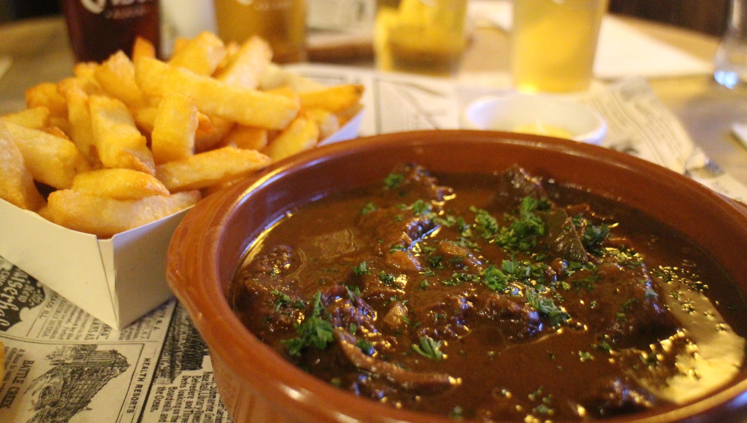 Pile of golden French fries beside a hearty beef stew in a brown ceramic bowl, garnished with parsley.