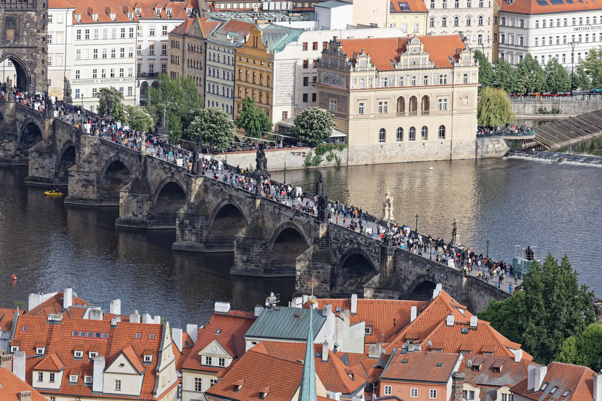 Crowded Charles Bridge across the Vltava River in Prague, with historic stone arches and red-tiled rooftops behinds.