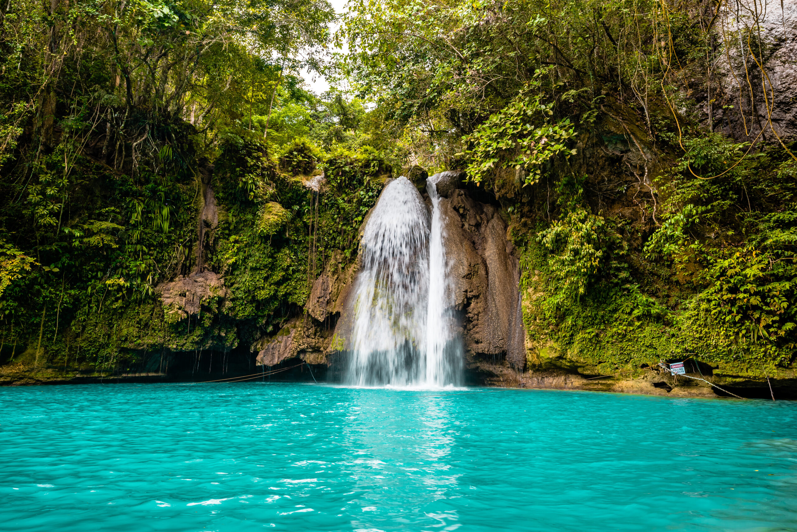 Waterfall drops from rocky ledge into a turquoise pool, surrounded by dense tropical jungle.