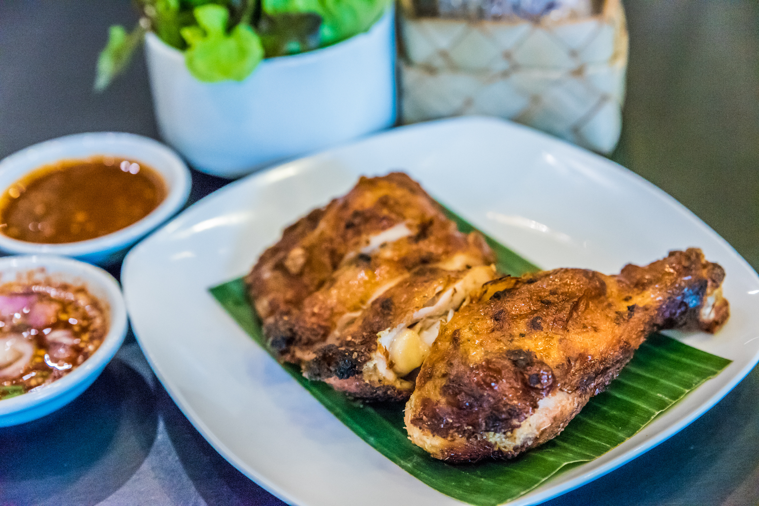 Plate with crispy fried chicken pieces served on a banana leaf, plus small bowls of dipping sauces nearby and a plant in the background.