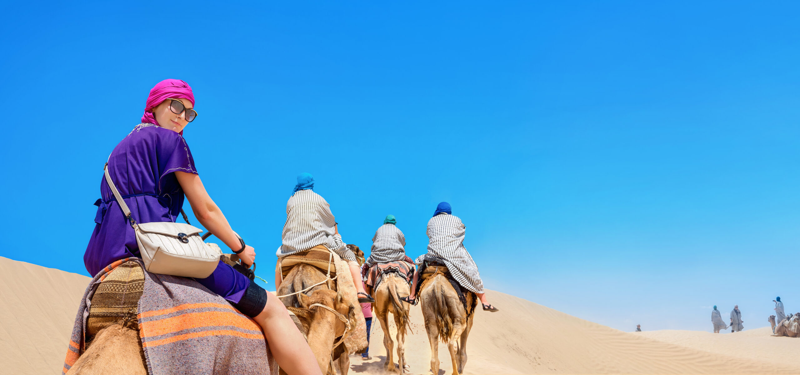 Group of people riding camels across a sandy desert under a bright blue sky in daytime.