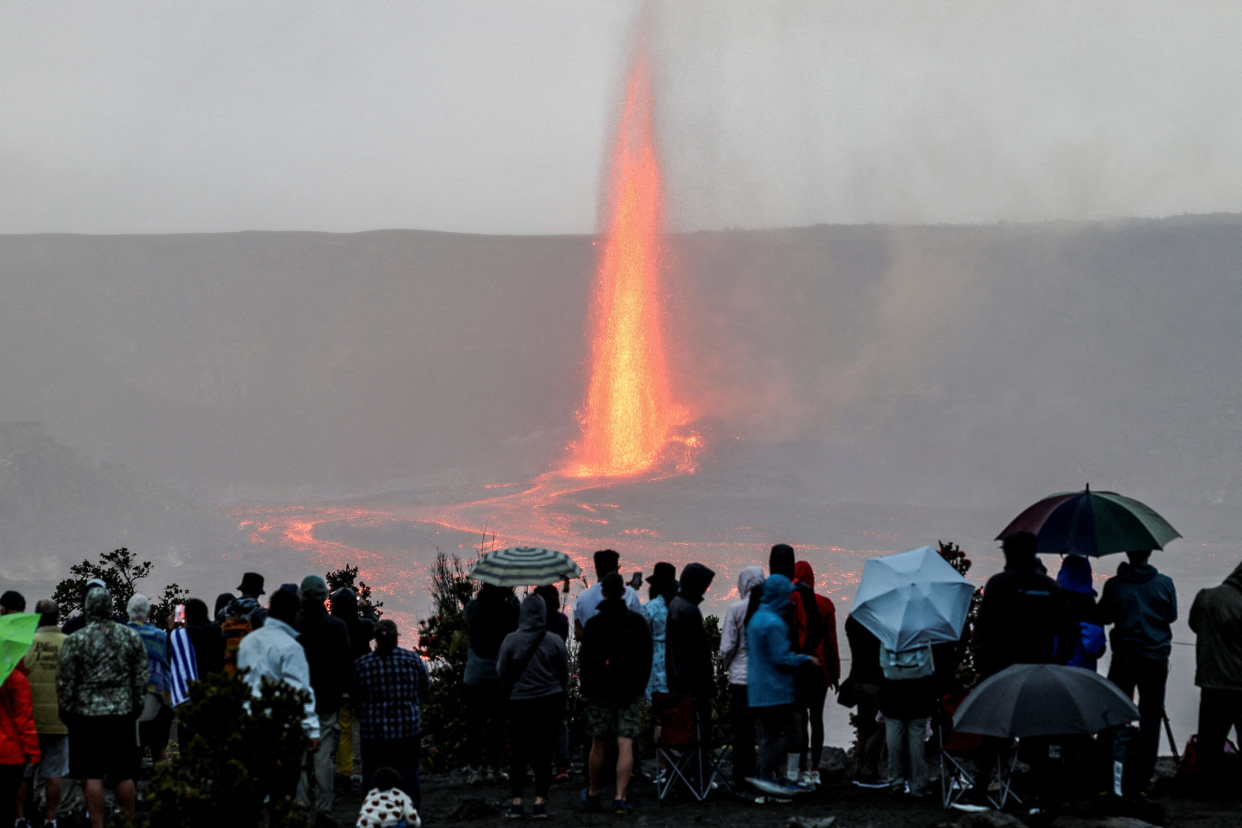 Crowd watching a lava fountain eruption at the shore, some umbrellas raised against the rain or mist.