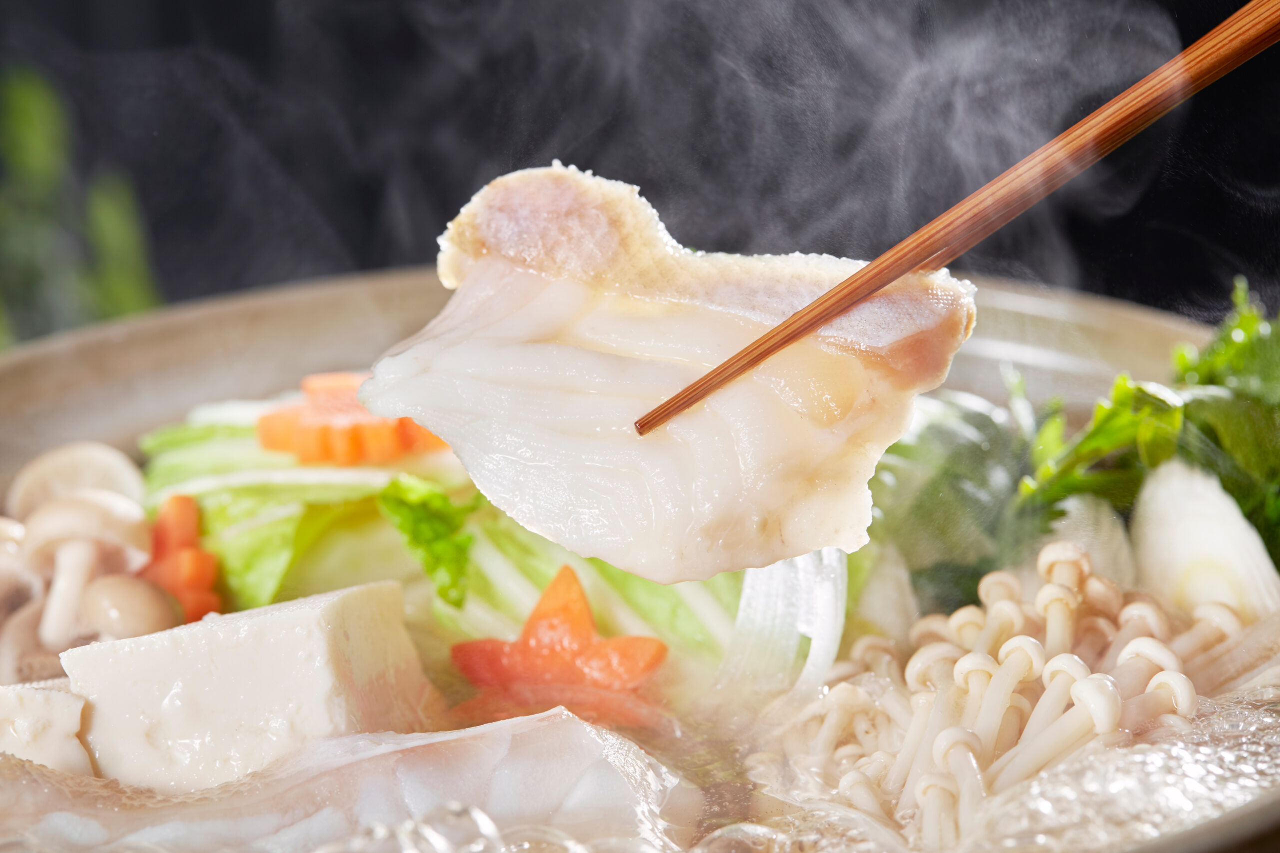 Slice of pork held by chopsticks above a steaming hot pot with tofu, mushrooms, and vegetables in the background