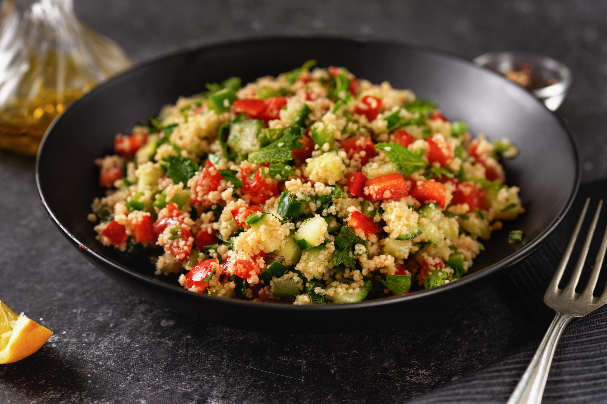 Couscous salad with tomatoes, cucumber and herbs in a black bowl on a dark counter