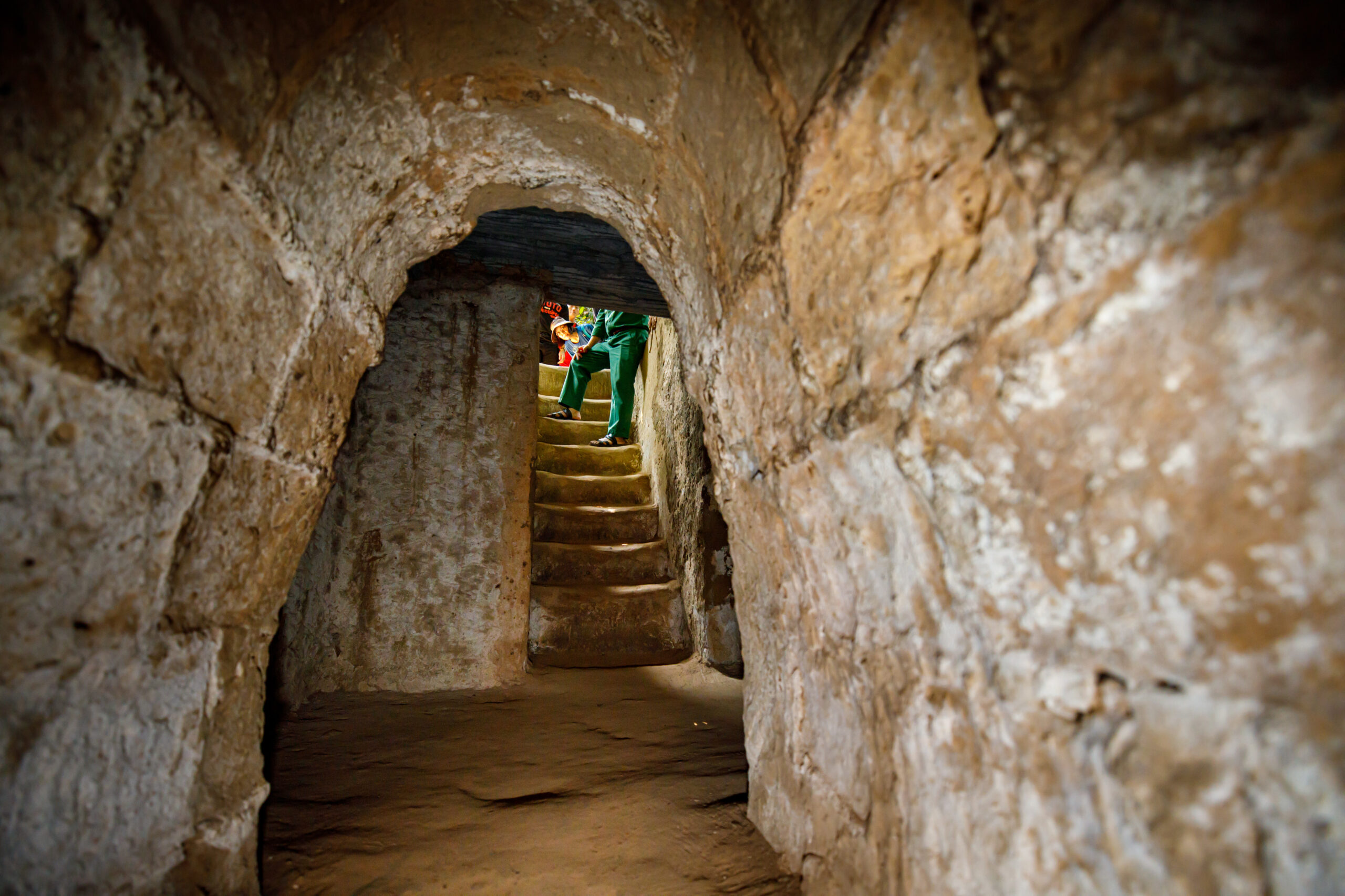 A person in green clothes climbing stone stairs inside a rocky cave opening, seen through a narrow tunnel.
