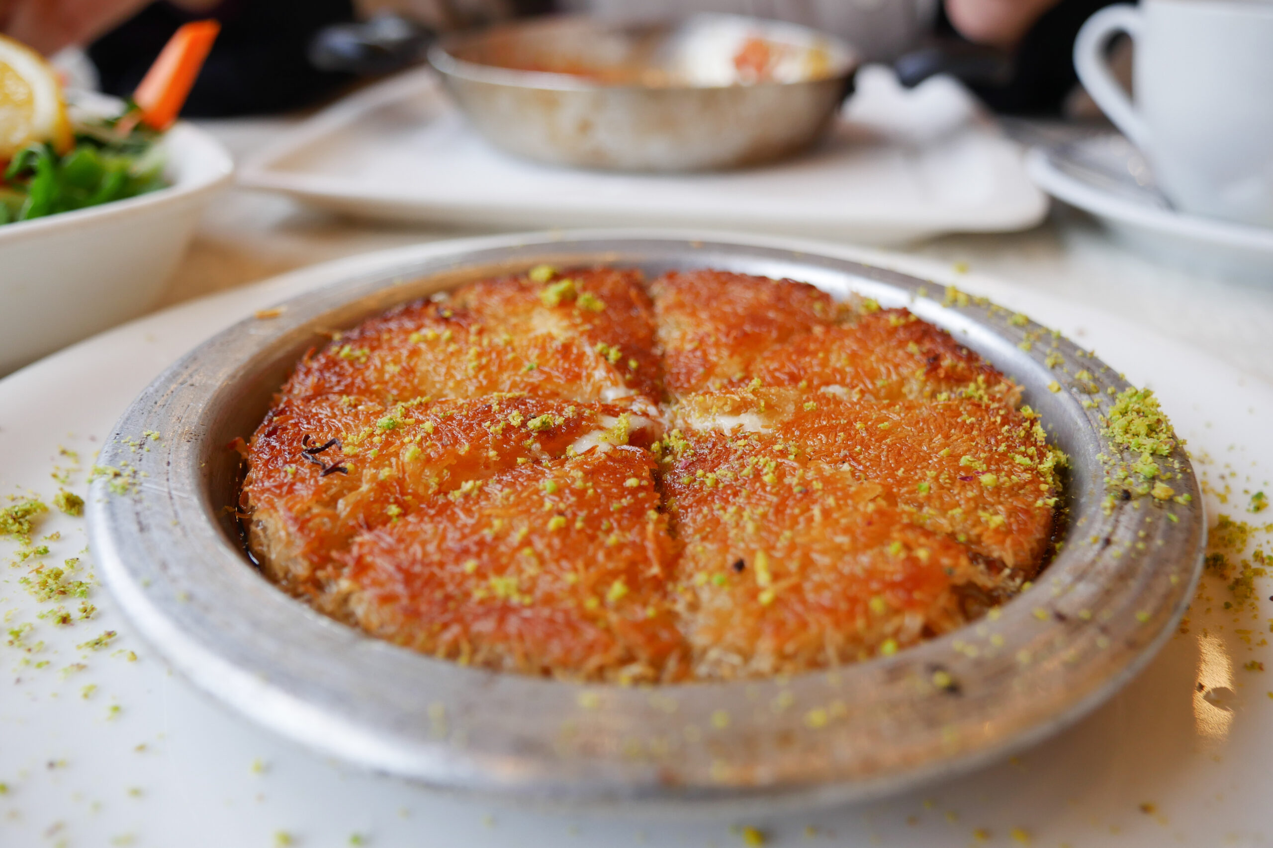 Baklava in a metal dish topped with pistachio sprinkles and orange glaze on a white plate.