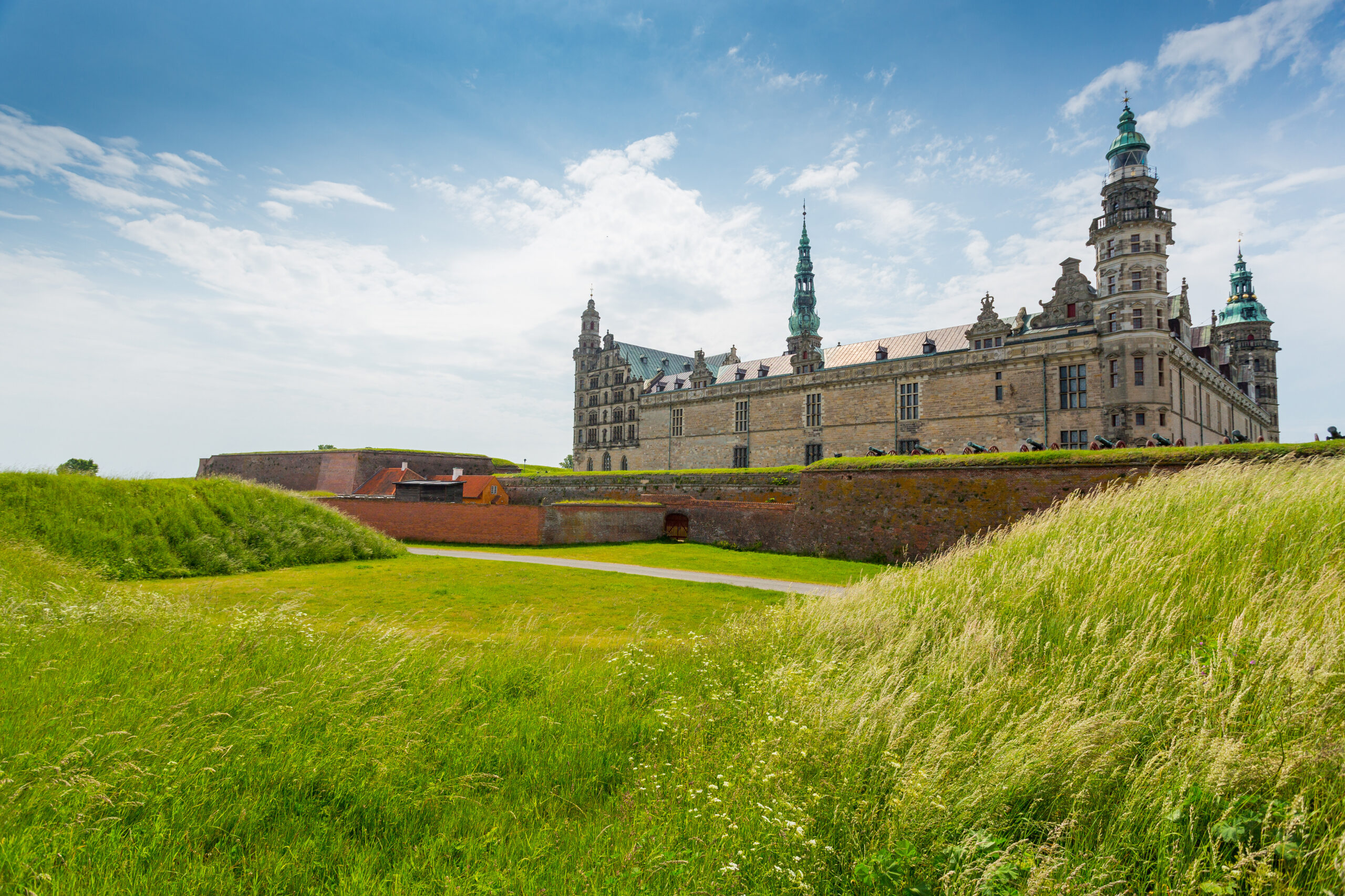 Stone castle with ornate towers and copper domes sits atop a brick rampart, under a bright blue sky with scattered clouds.