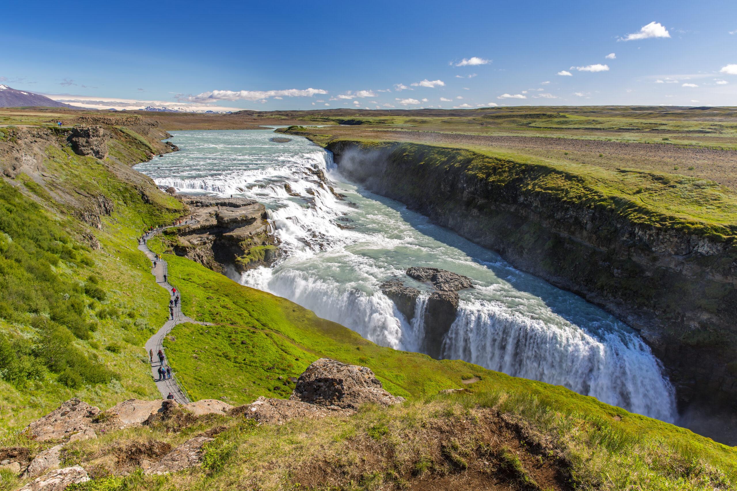 Wide Icelandic canyon with a powerful waterfall and turquoise river, green slopes, and a winding boardwalk with hikers under a bright blue sky