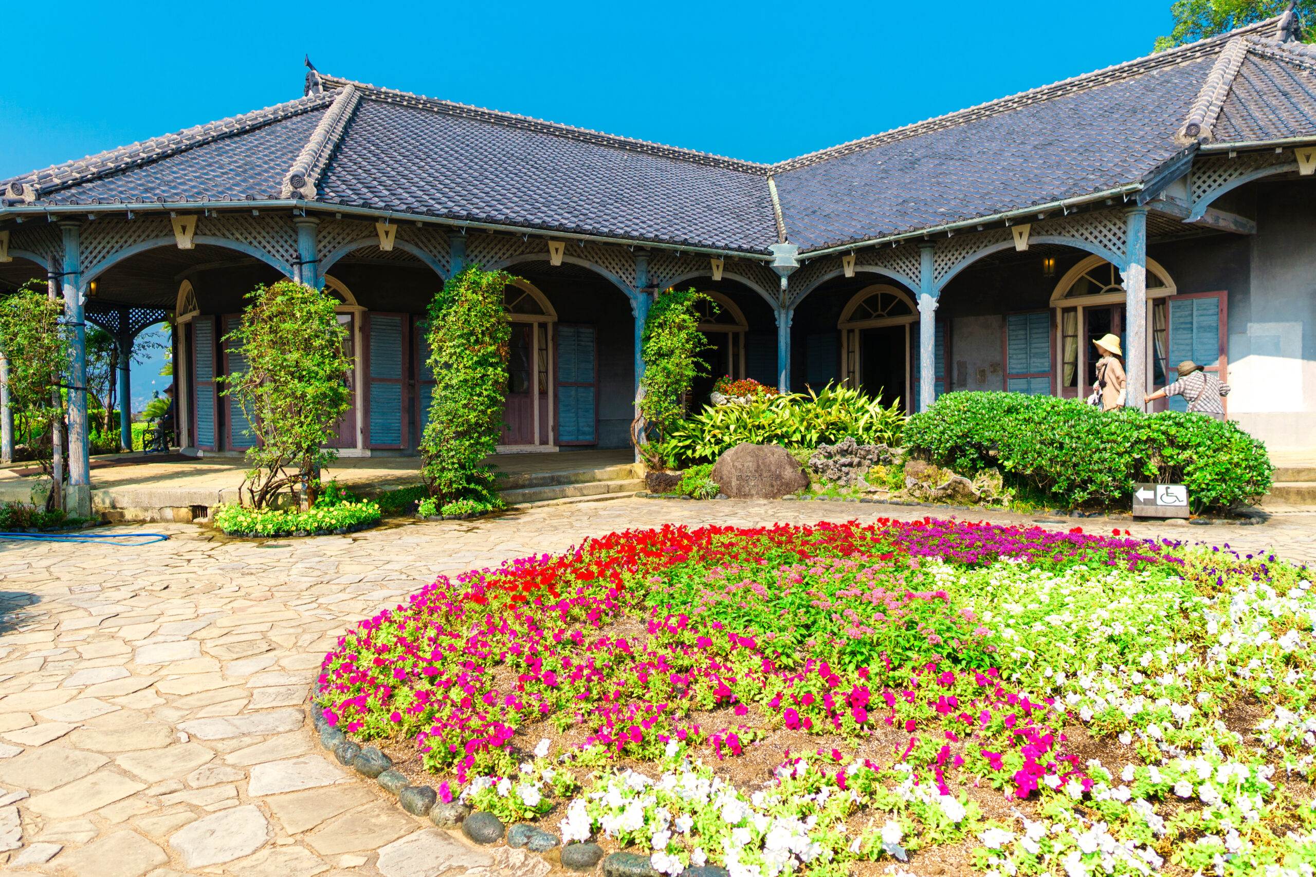 Courtyard with a blue-trimmed veranda and tiled roof, colorful flower beds along a stone path, and visitors on the right side.
