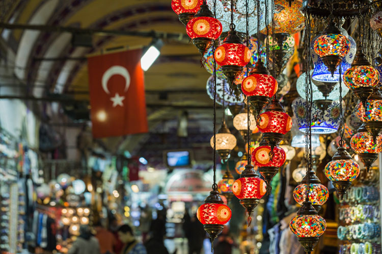 Colorful mosaic lanterns hanging in a Turkish bazaar with a Turkish flag in the background, shoppers blurred in the distance.