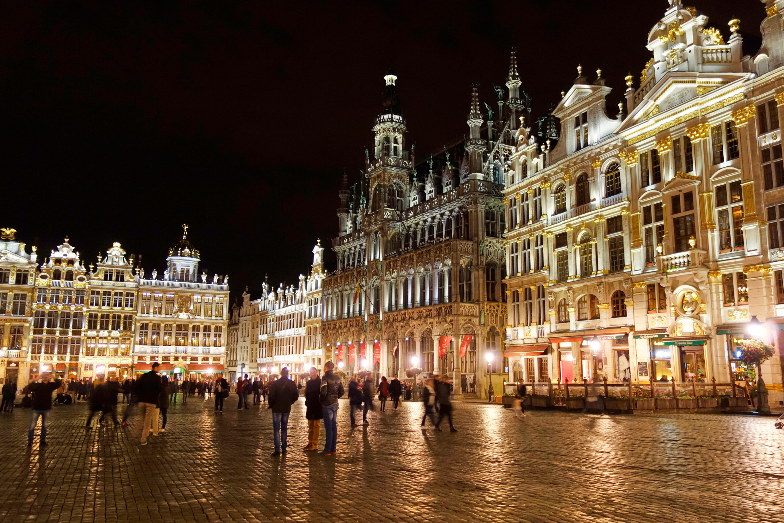 Night view of a historic square with ornate gold-trimmed buildings and crowds walking on a cobblestone plaza. (Brussels' Grand Place at night)