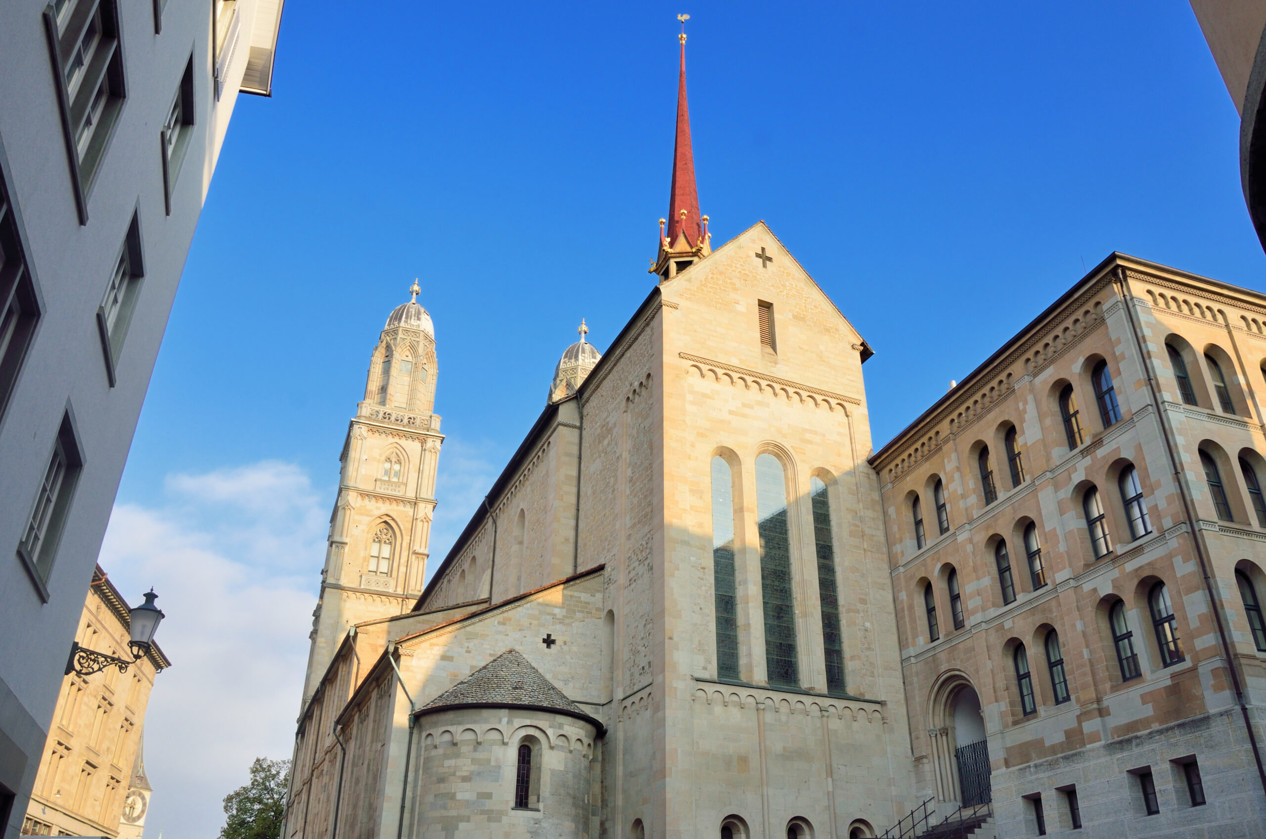 Stone church with a tall red spire and cross, set against a bright blue sky, flanked by historic beige buildings and towers on a sunny day