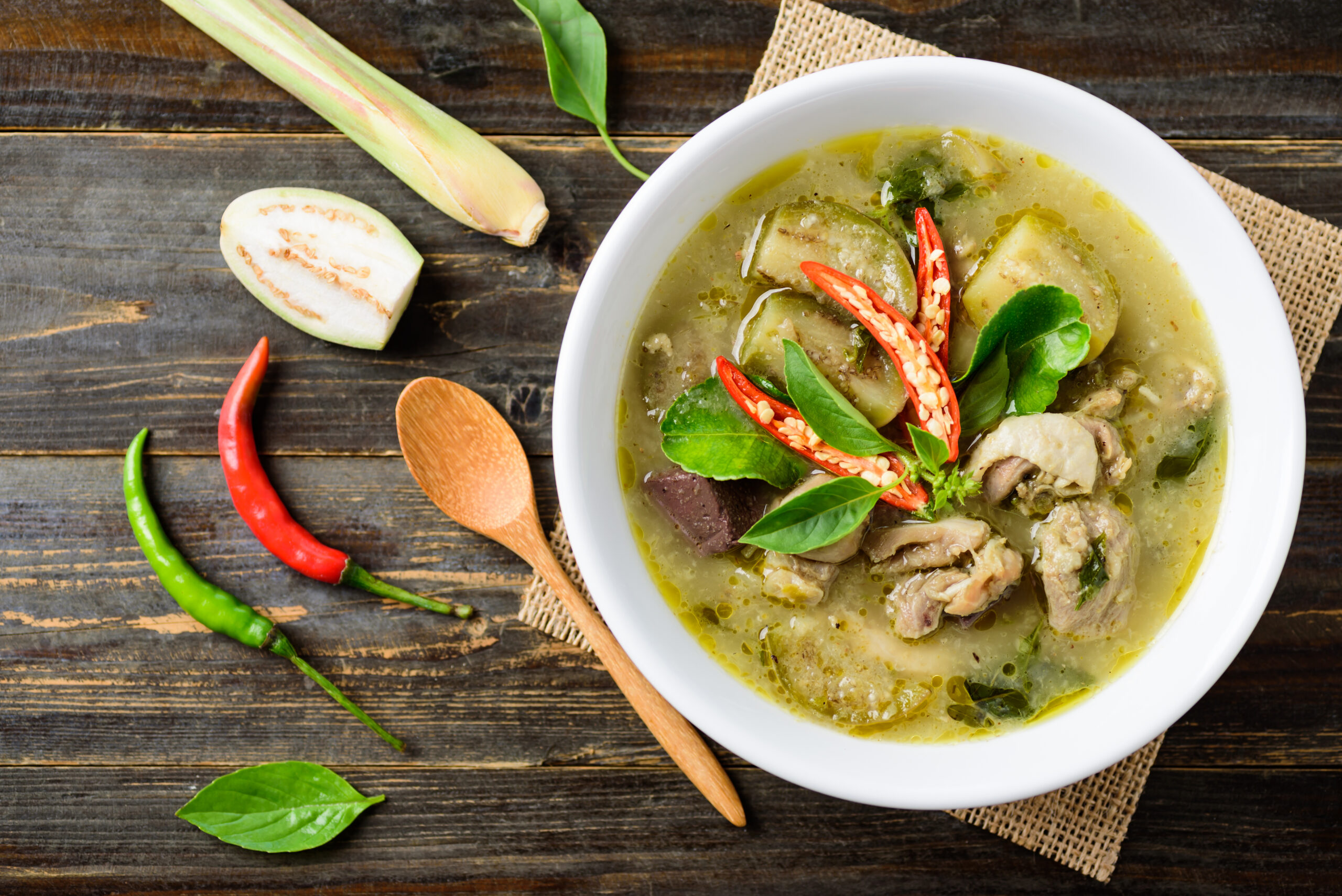 White bowl of green curry soup with eggplant, basil and red chili garnish on a dark wooden table, with a wooden spoon nearby.