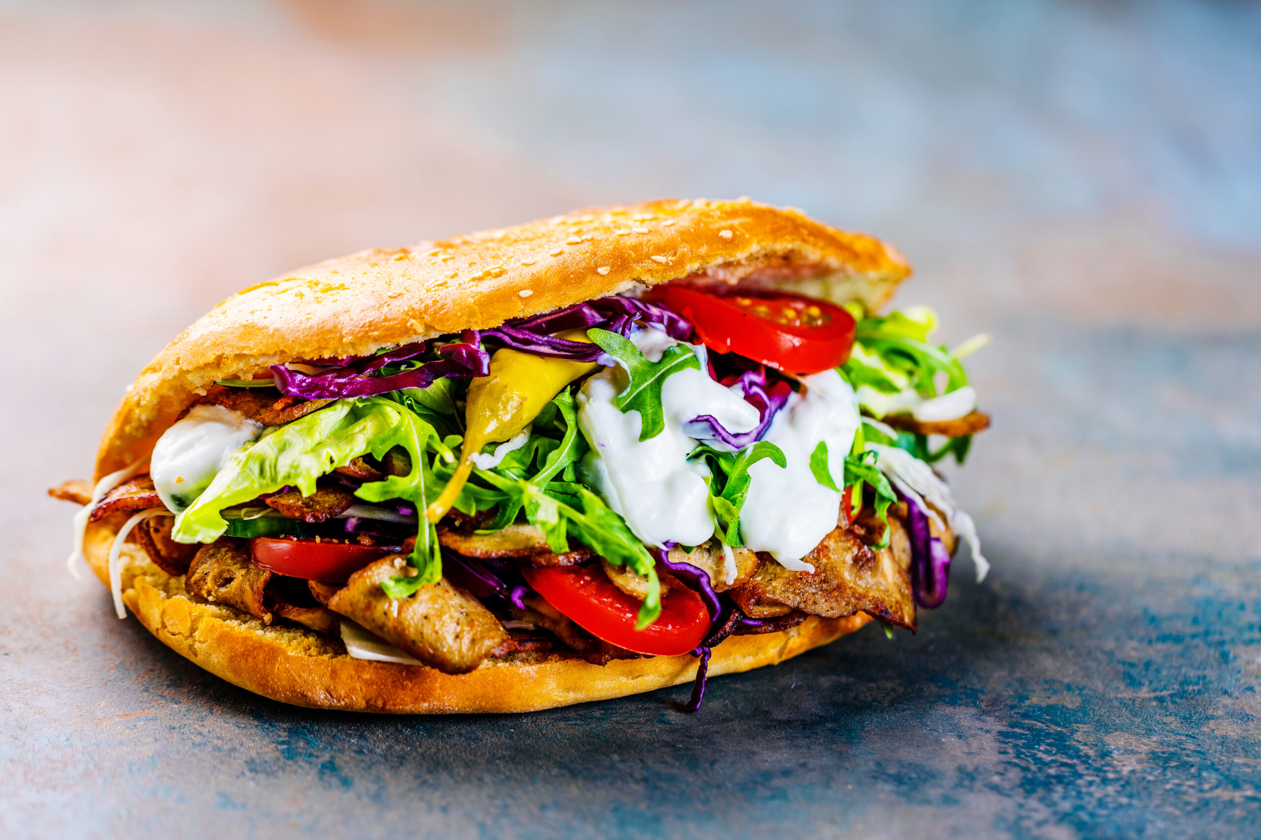 Close-up of a loaded sesame-bun burger with lettuce, tomato, red cabbage, greens, and creamy sauce on top.