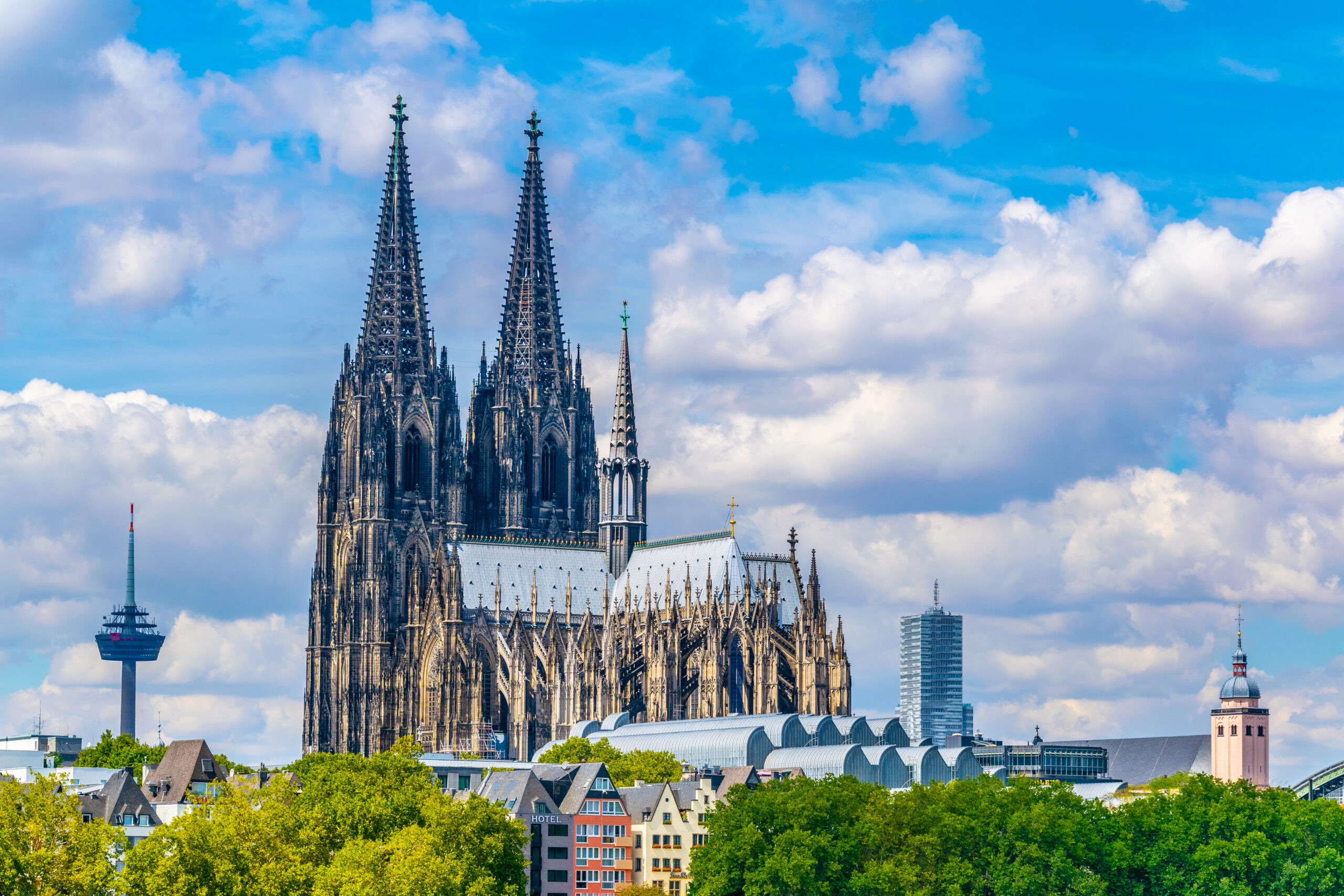 Cologne Cathedral's Gothic spires rise above a cityscape with trees and modern buildings under a blue sky.