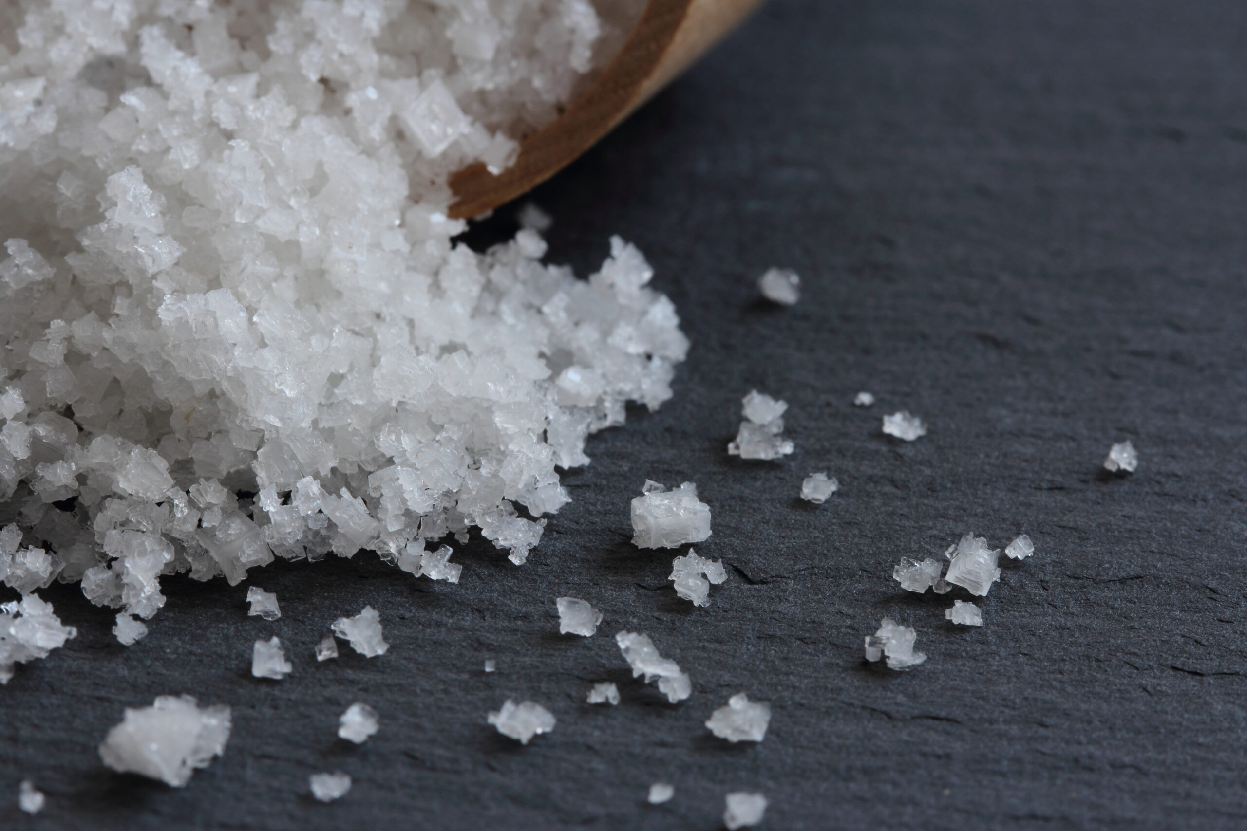 Coarse white salt crystals spilled from a wooden scoop onto a dark slate surface.