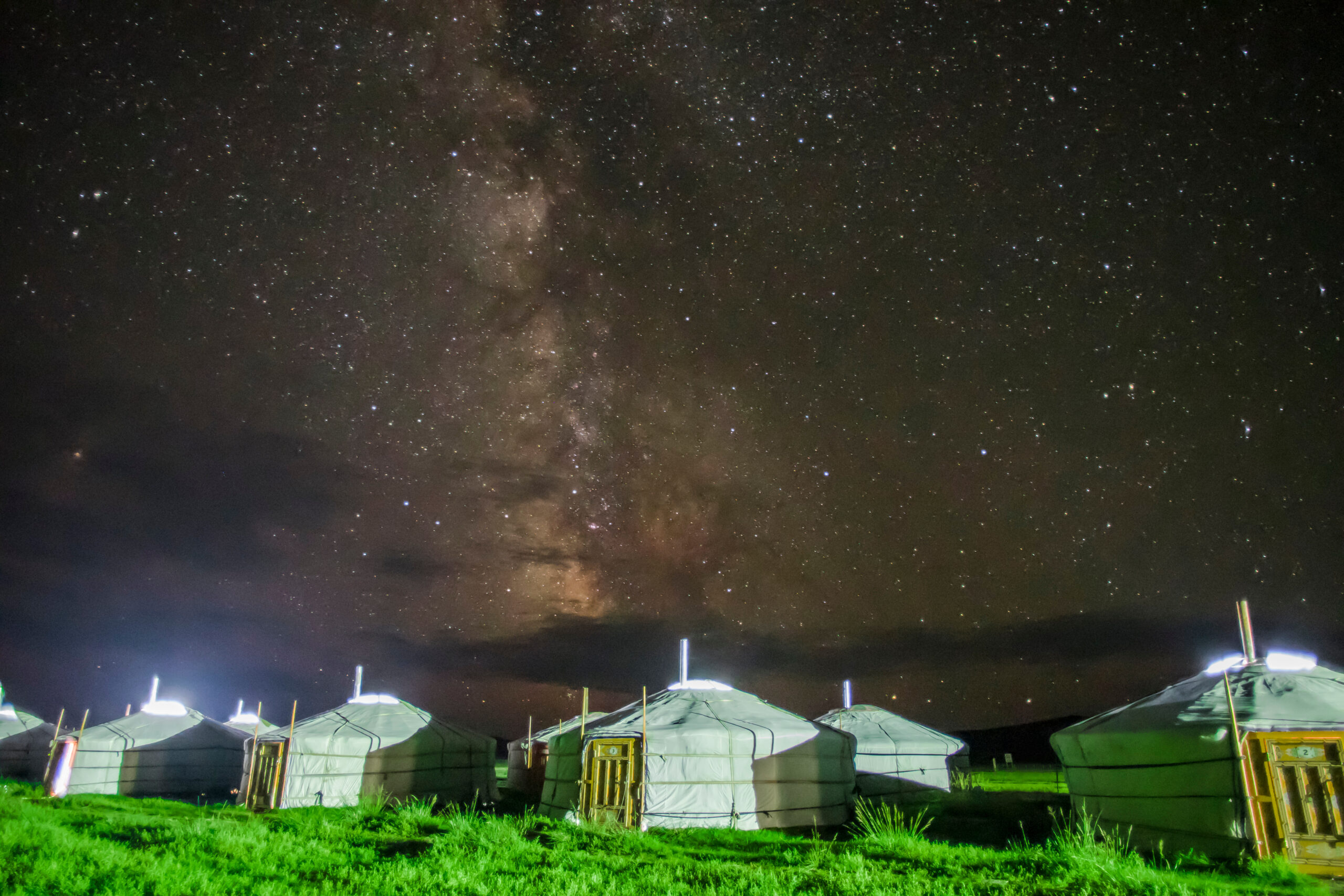 Night sky full of stars and the Milky Way above a row of white yurts on a grassy field at night.