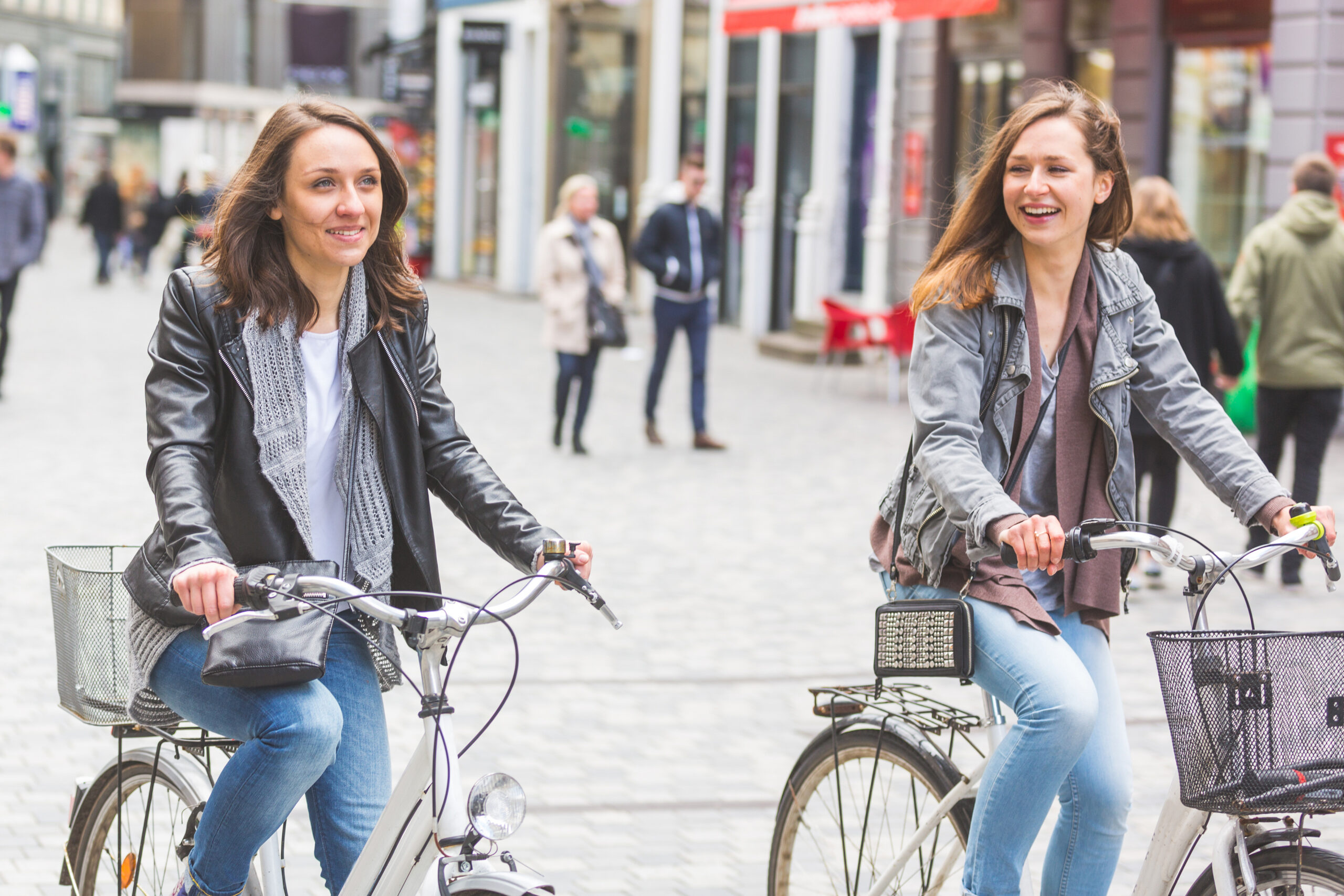 Two young women ride bicycles on a cobblestone city street, smiling and chatting in jackets and scarves in the foreground.