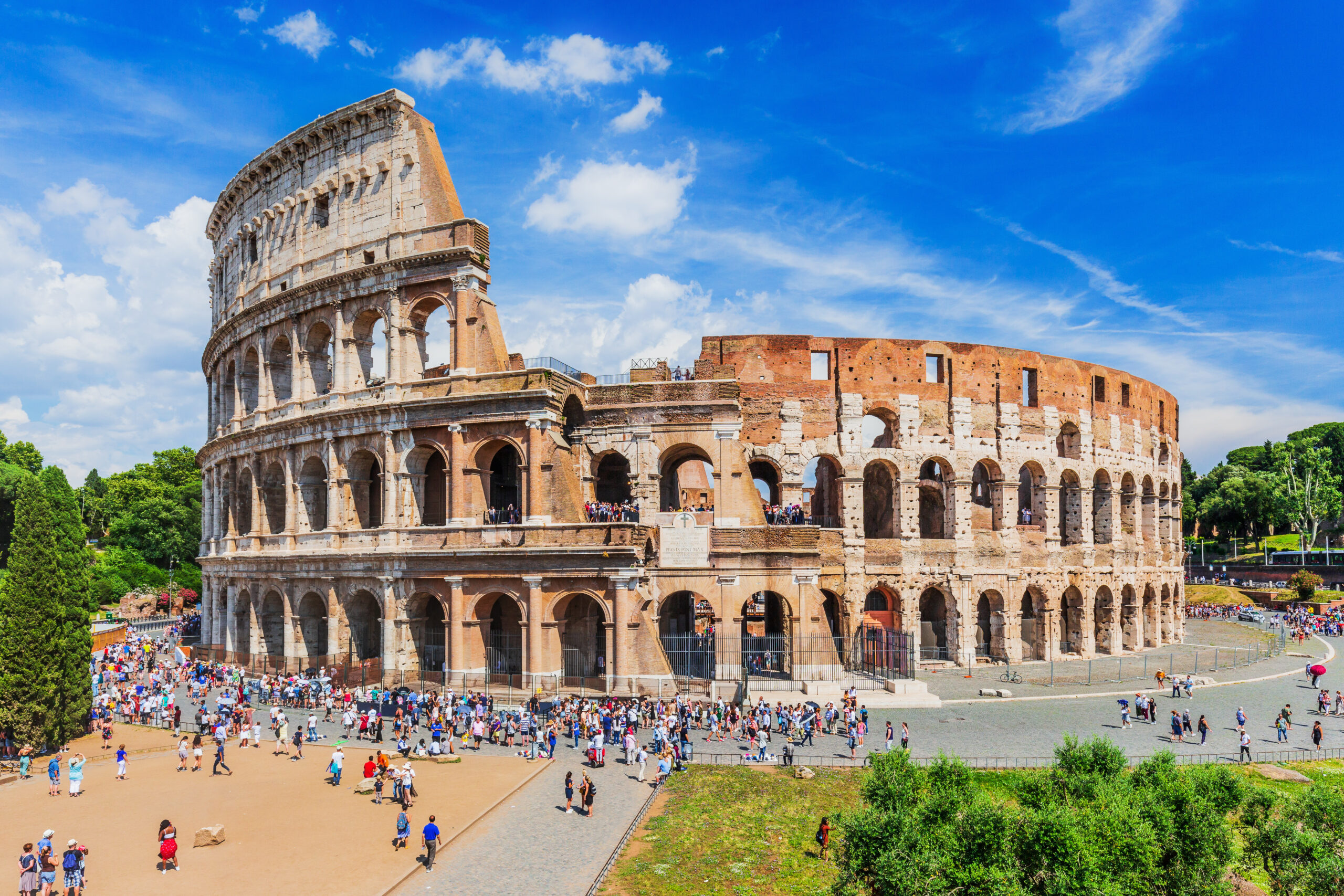 Exterior view of the Roman Colosseum with arched colonnades and a crowd of tourists against a bright blue sky.