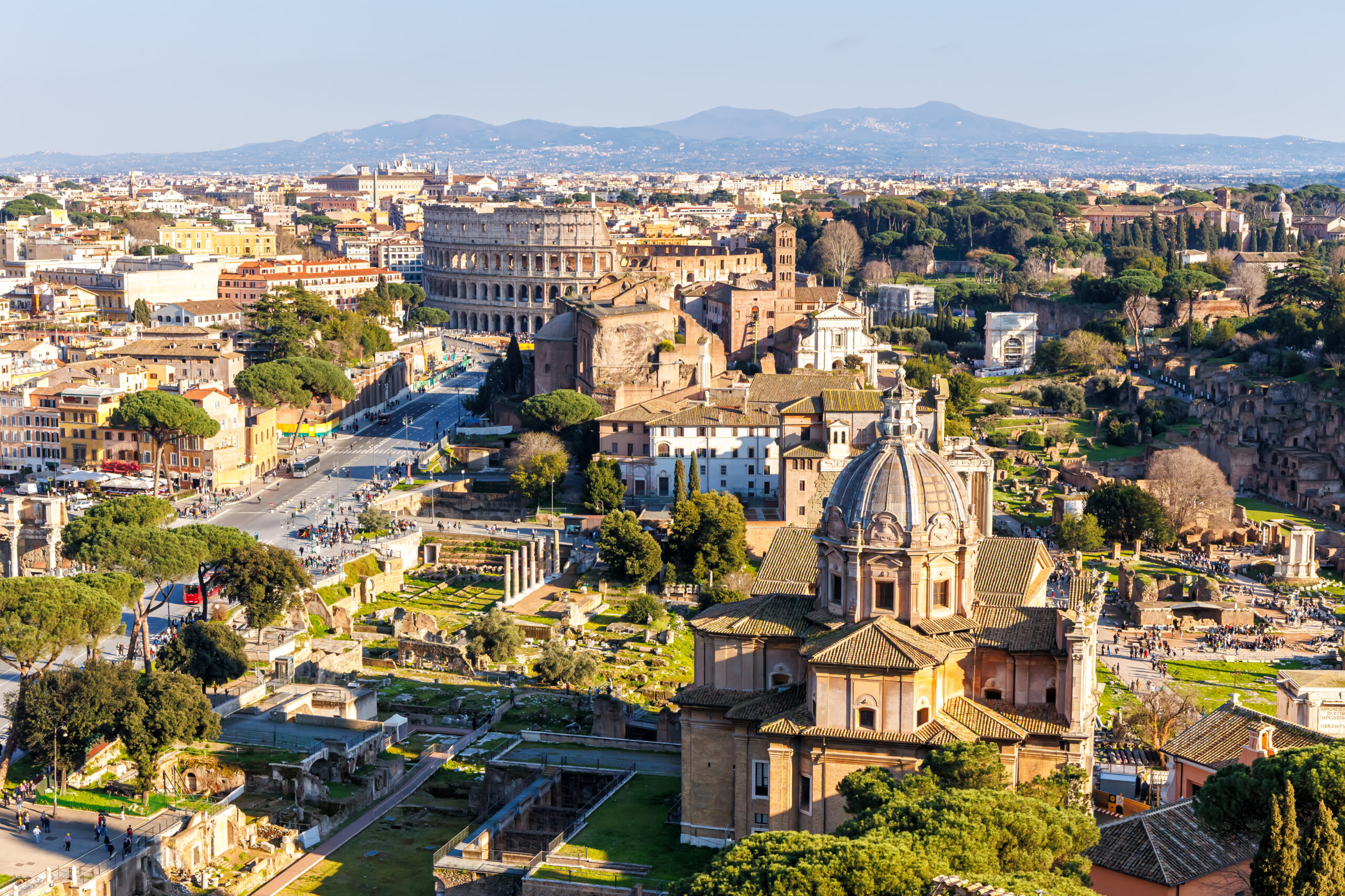 Aerial view of Rome showing the Colosseum, surrounding ruins, and city streets with greenery under a clear blue sky.