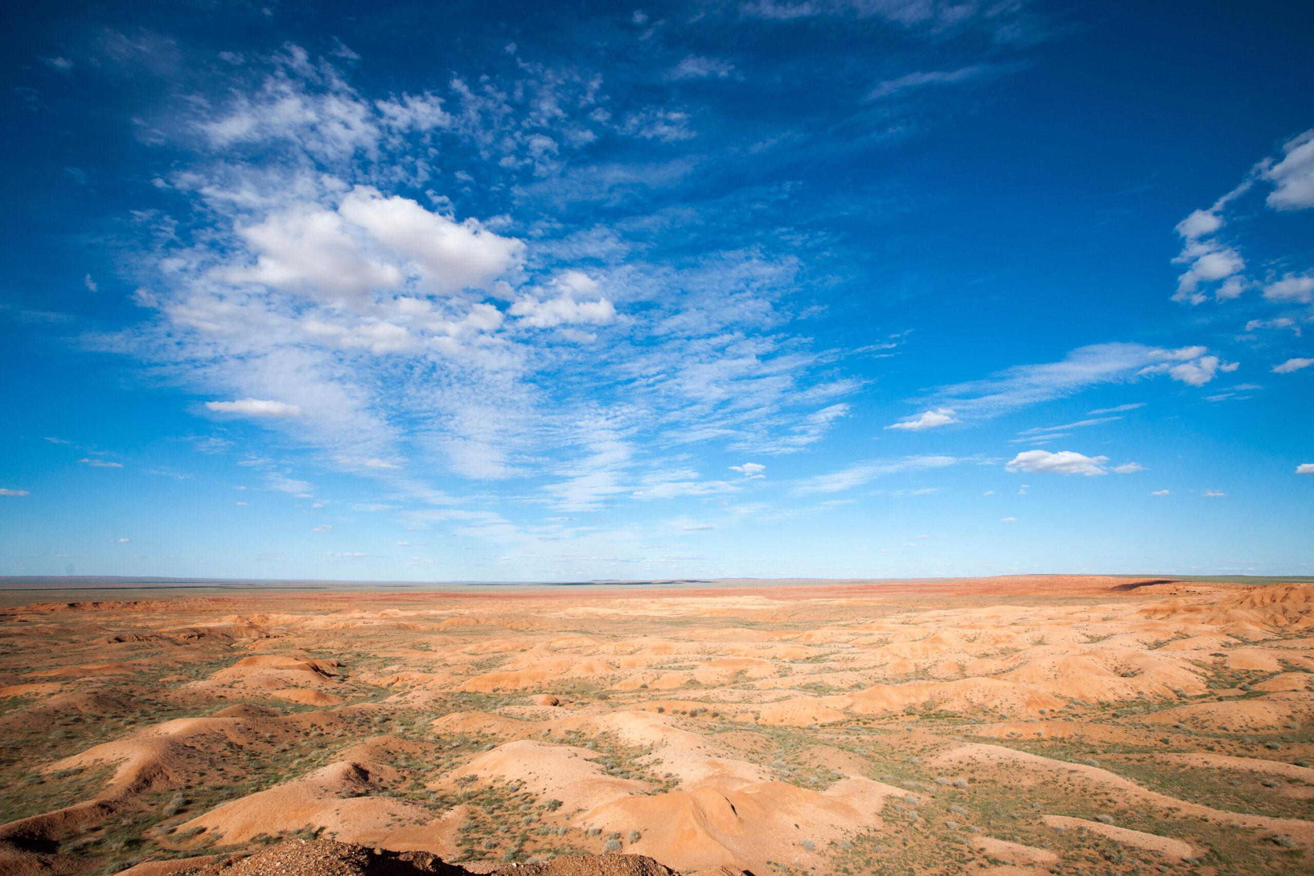 Wide desert landscape with orange sand dunes under a bright blue sky with scattered clouds.
