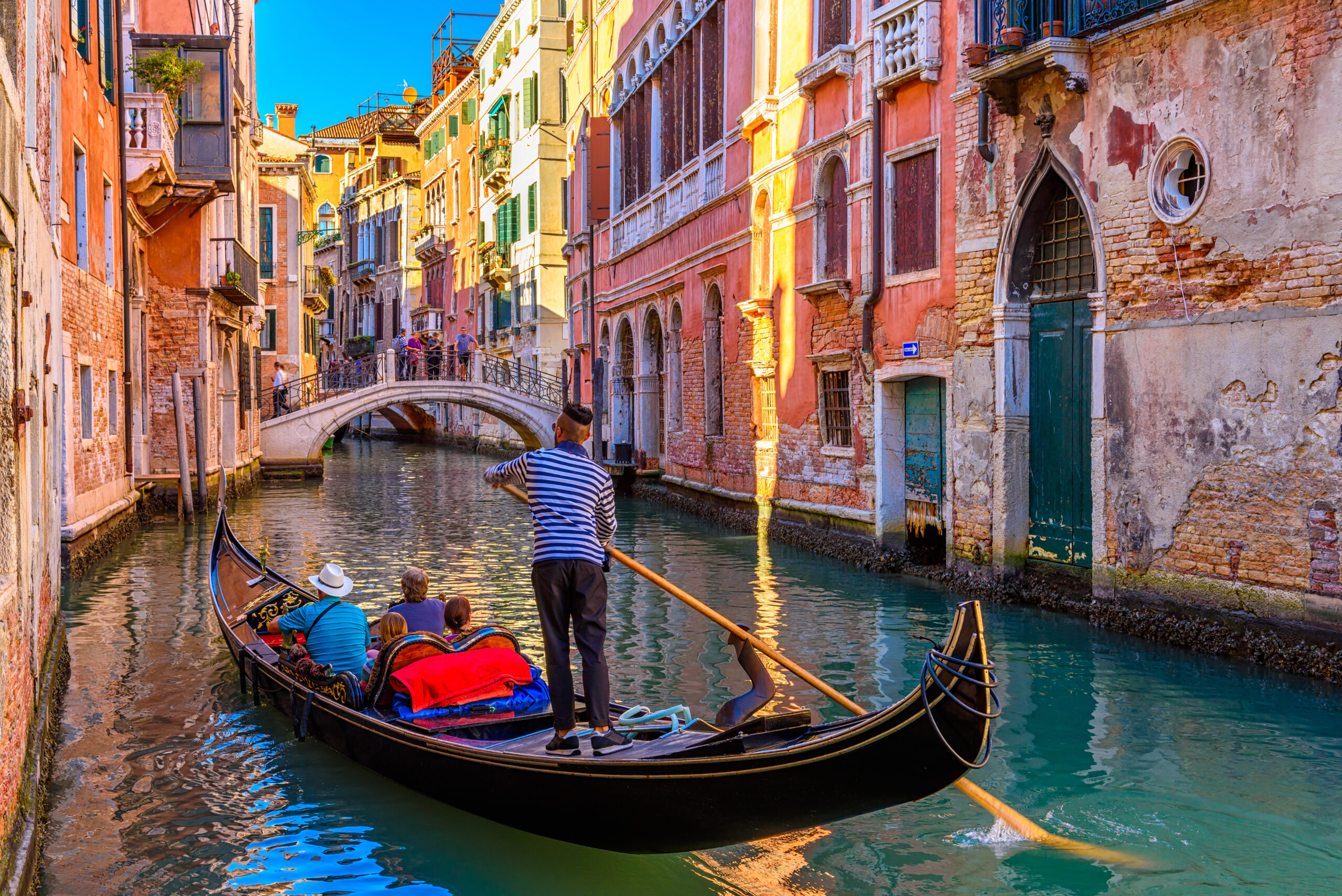Gondola carrying tourists gliding through a narrow canal in Venice, with colorful historic buildings and a small bridge ahead on a sunny day.