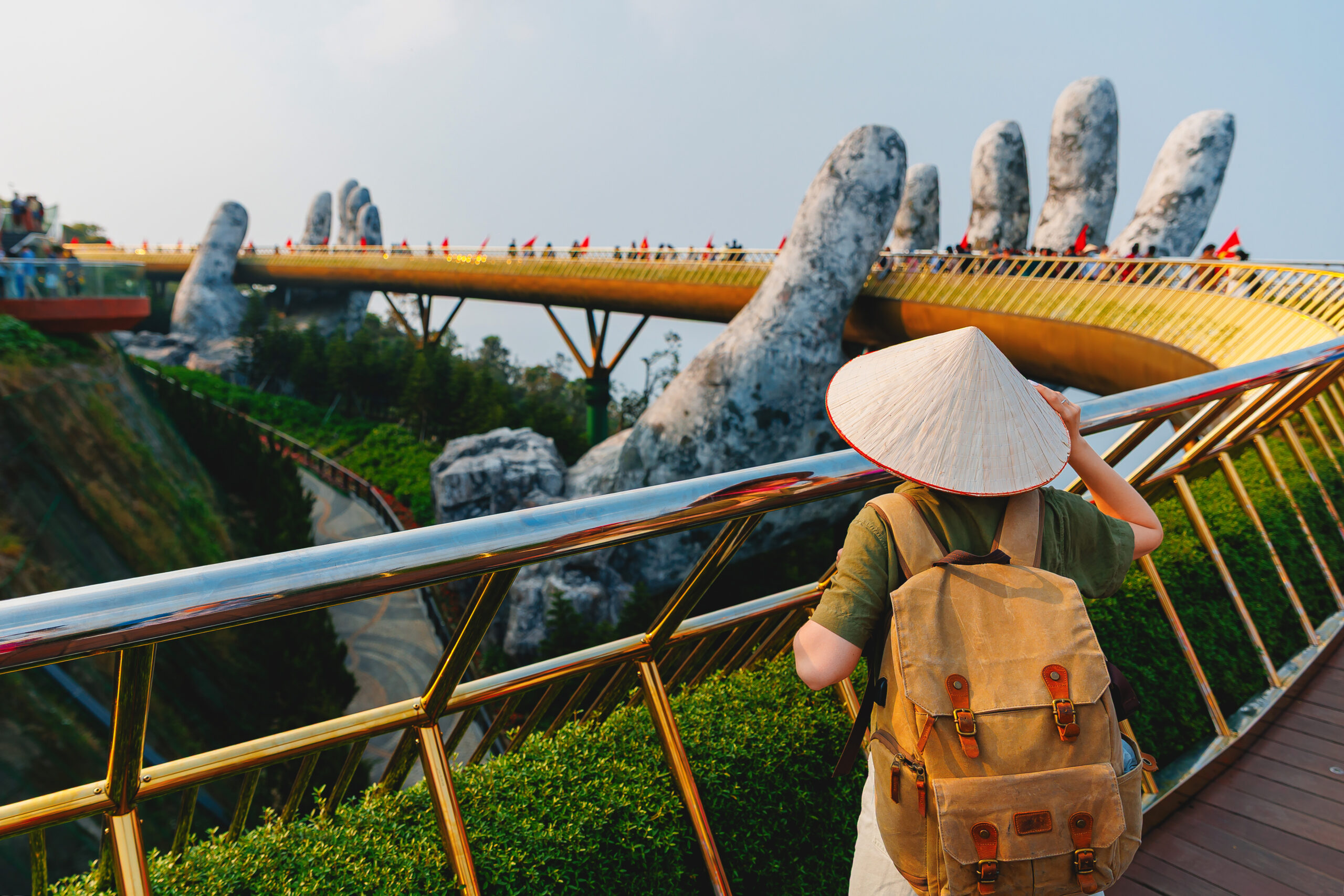 Person wearing a conical hat and backpack walks along a curved golden pedestrian bridge with giant finger-shaped pillars in the background.