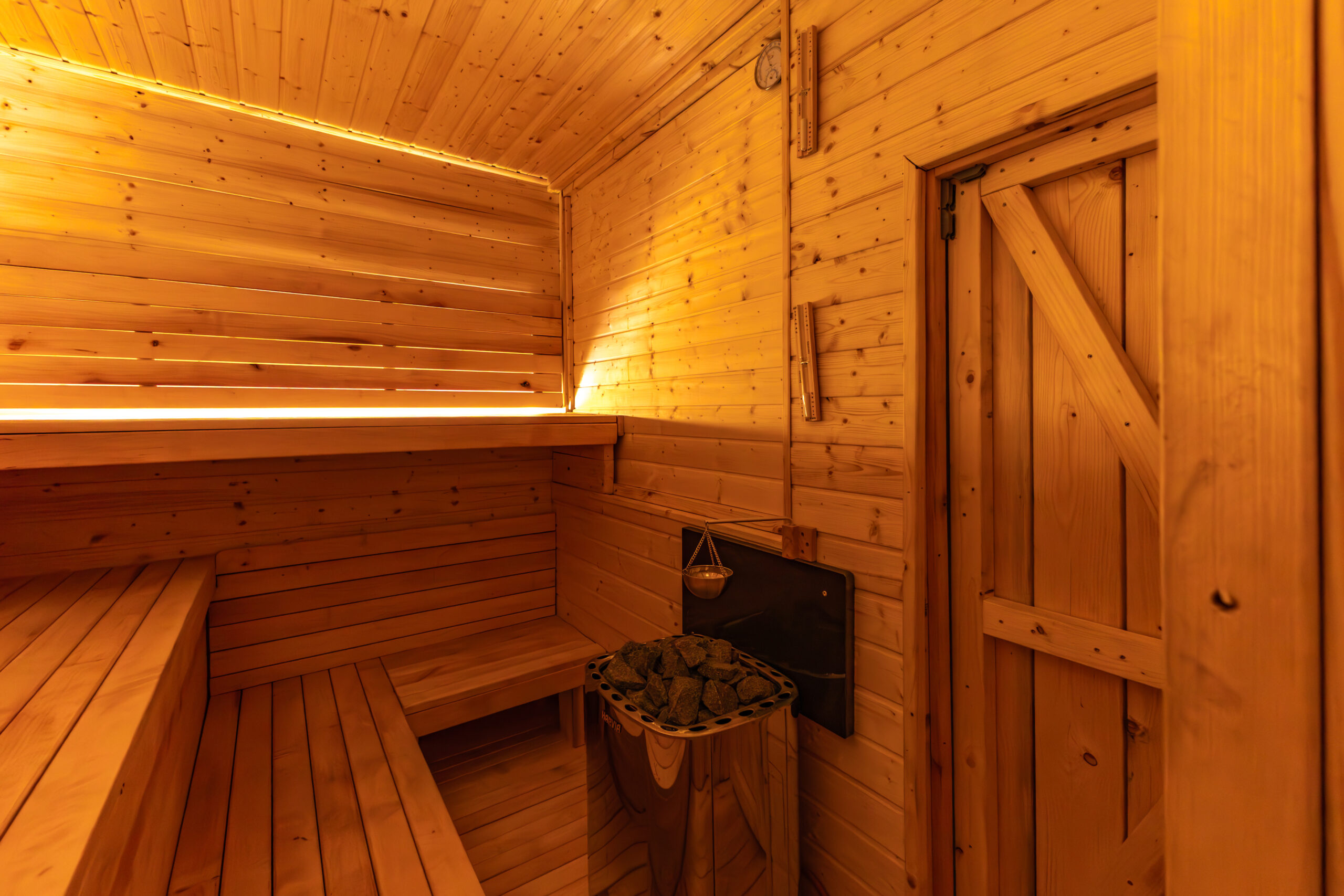 Wood-paneled sauna interior with tiered benches and a heater filled with sauna rocks nearby, warm amber lighting.