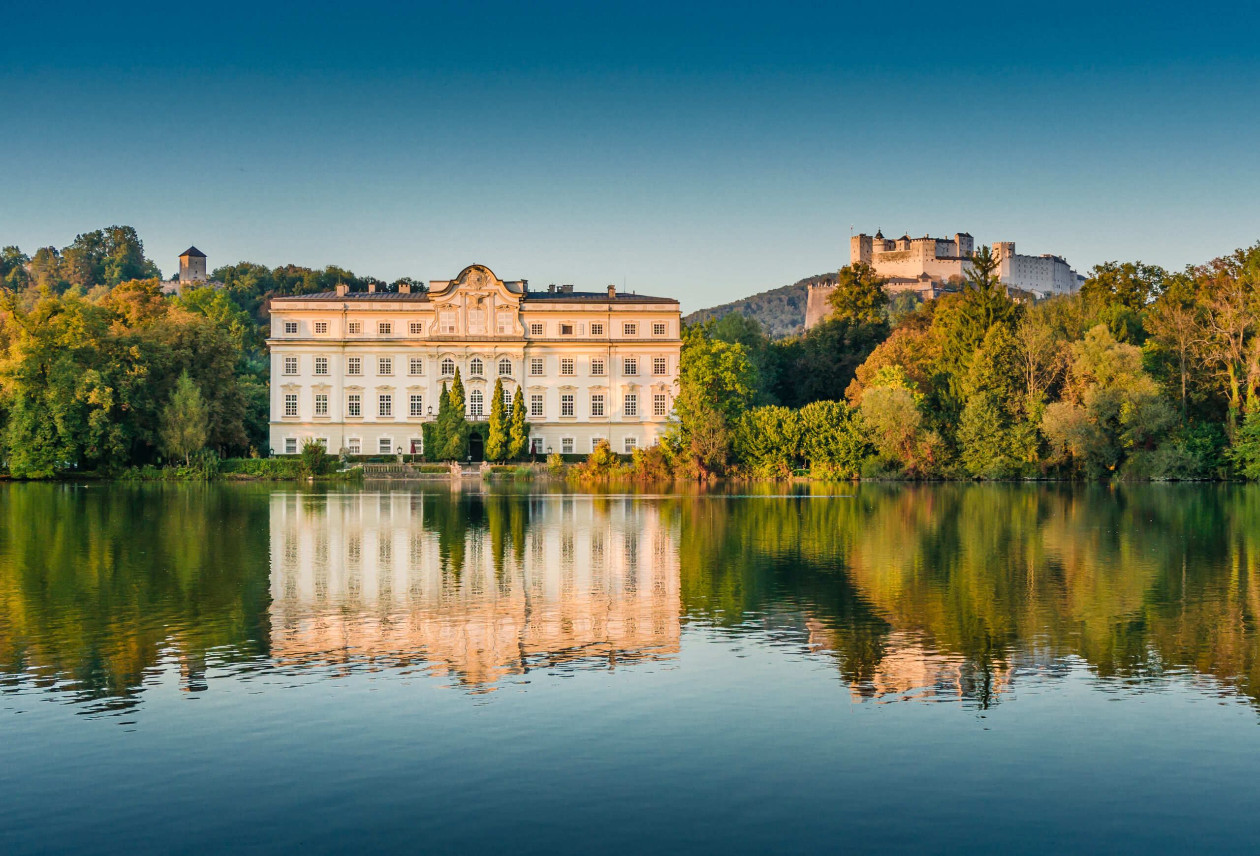 White historic building by a calm lake with a forested shore and a hilltop castle in the distance under a clear blue sky, reflects in the water