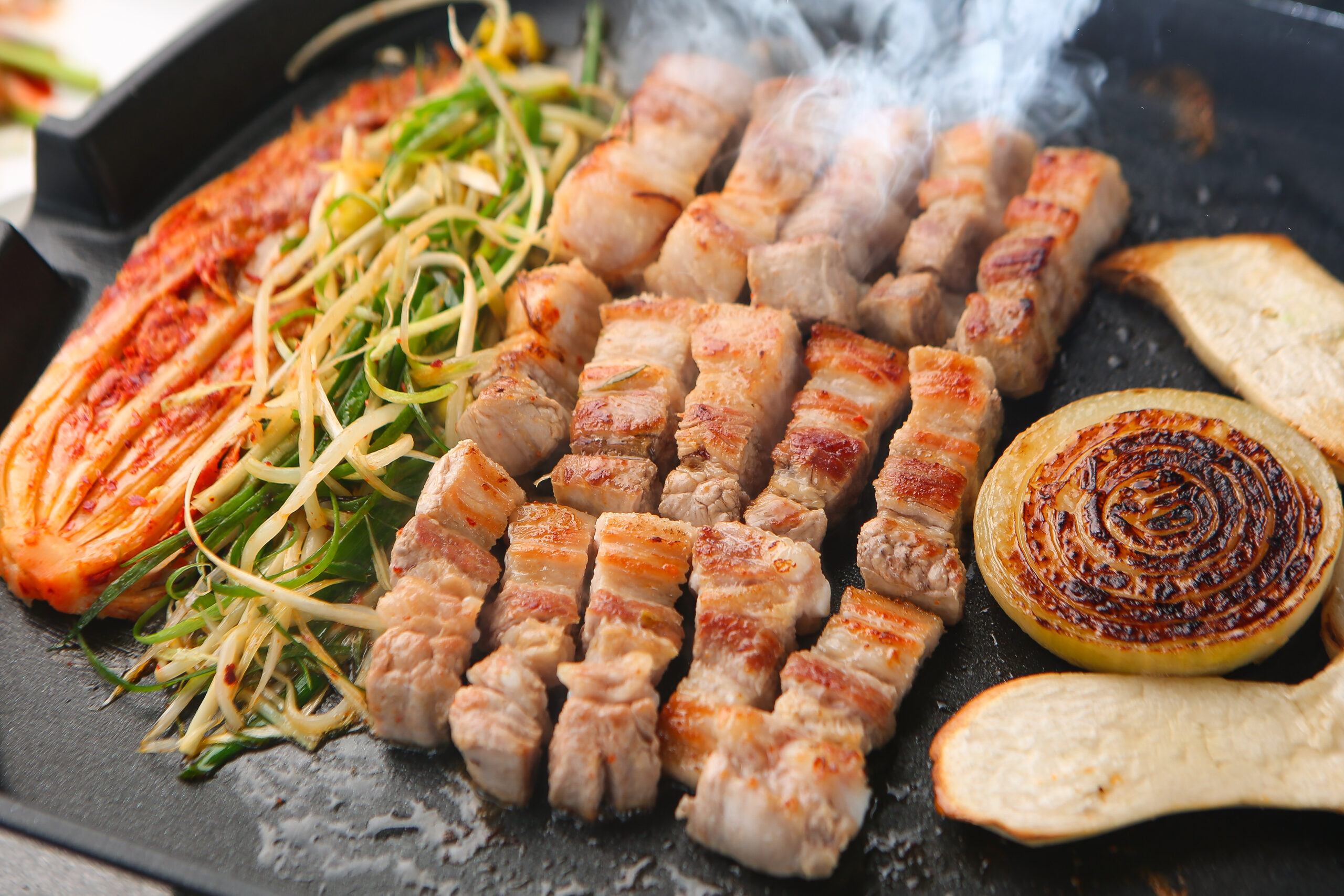 Grilled pork cubes, charred vegetables, and sliced onions on a hot skillet with greens and flatbread rounds nearby