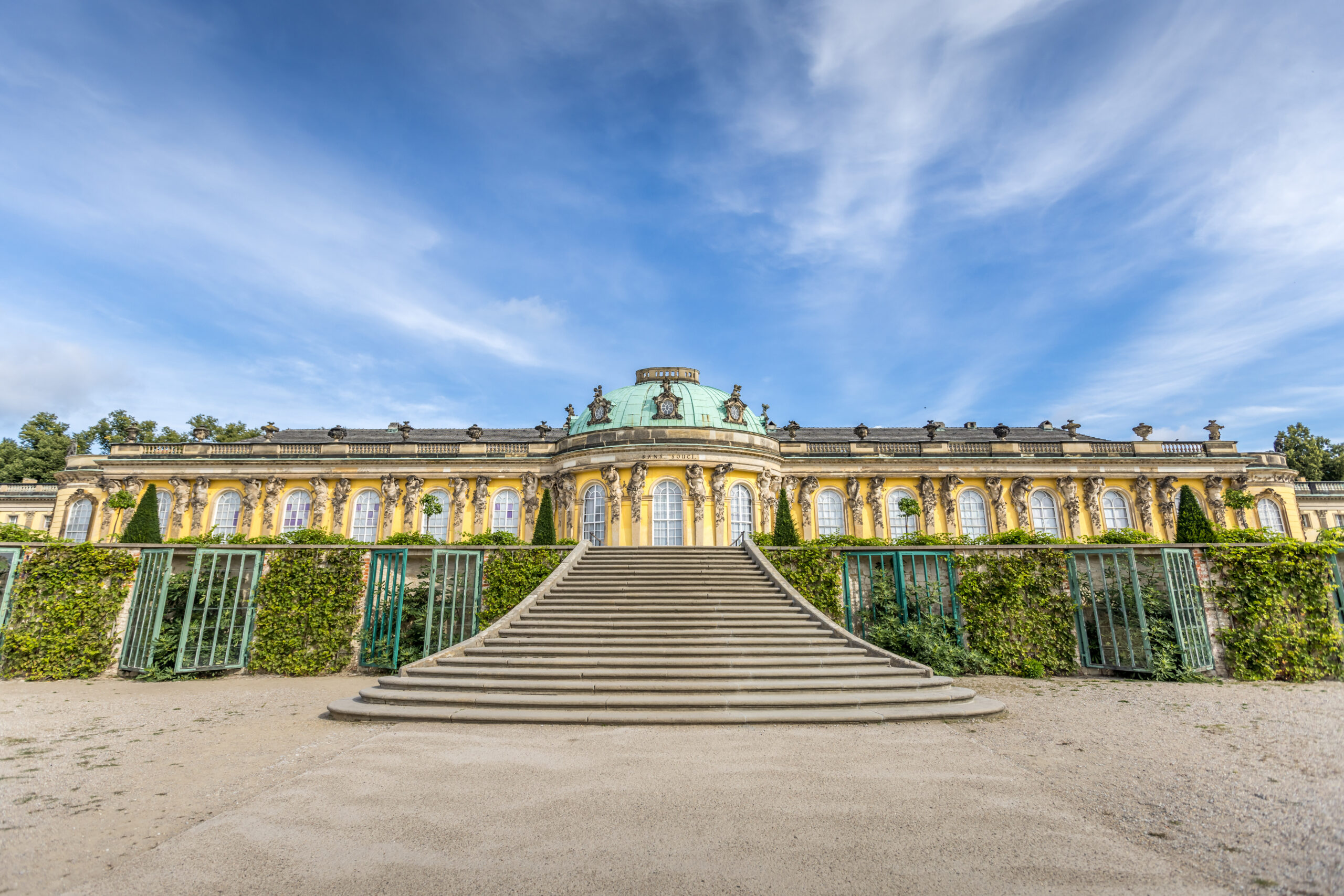 Front view of a yellow Baroque palace with a green domed roof, ornate statues along the balustrade, and a grand staircase reaching the entrance against a blue sky.