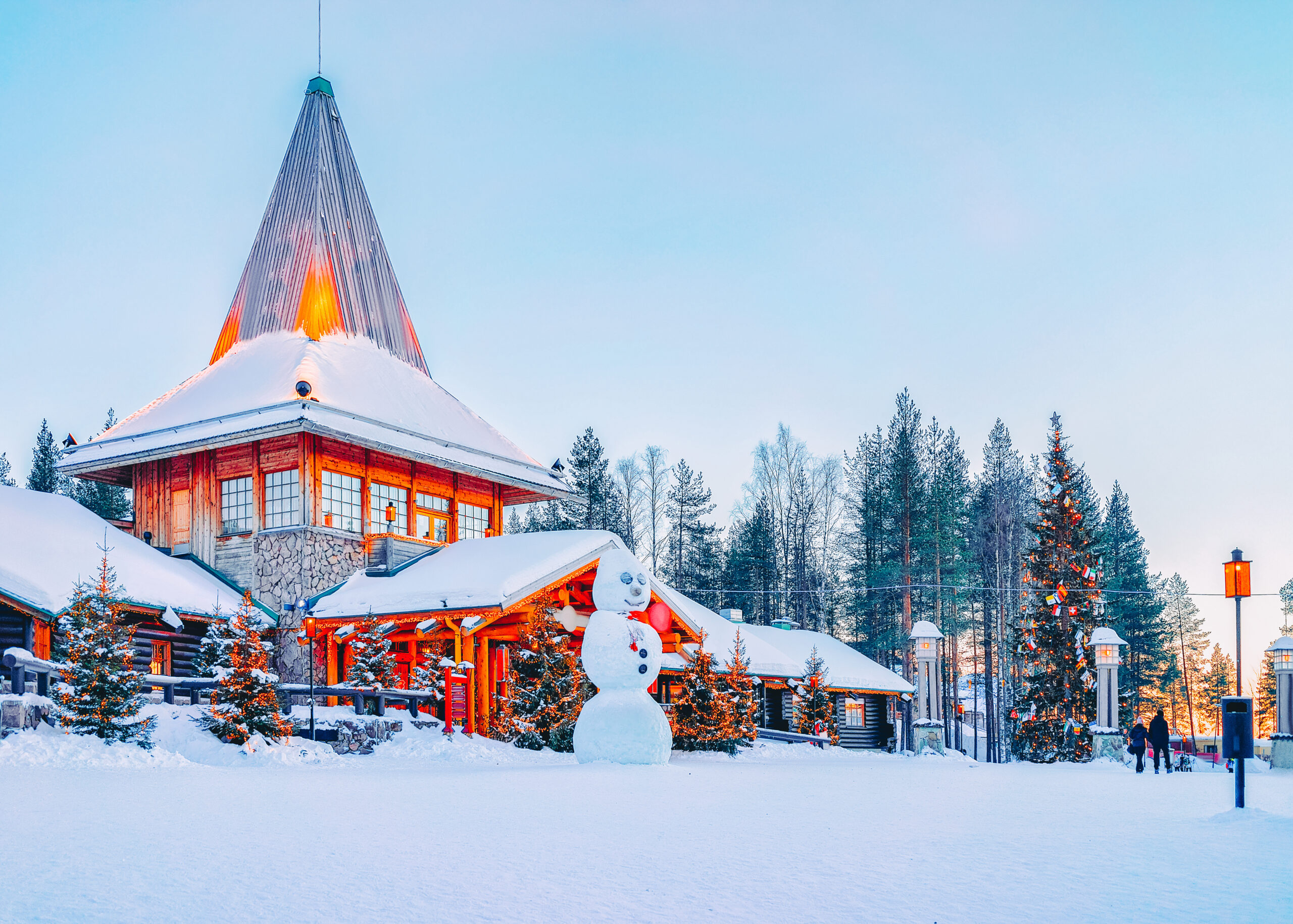 Snowy village scene with a wooden building and tall church steeple, snowman in foreground, and pine trees with Christmas lights under a pale blue sky.