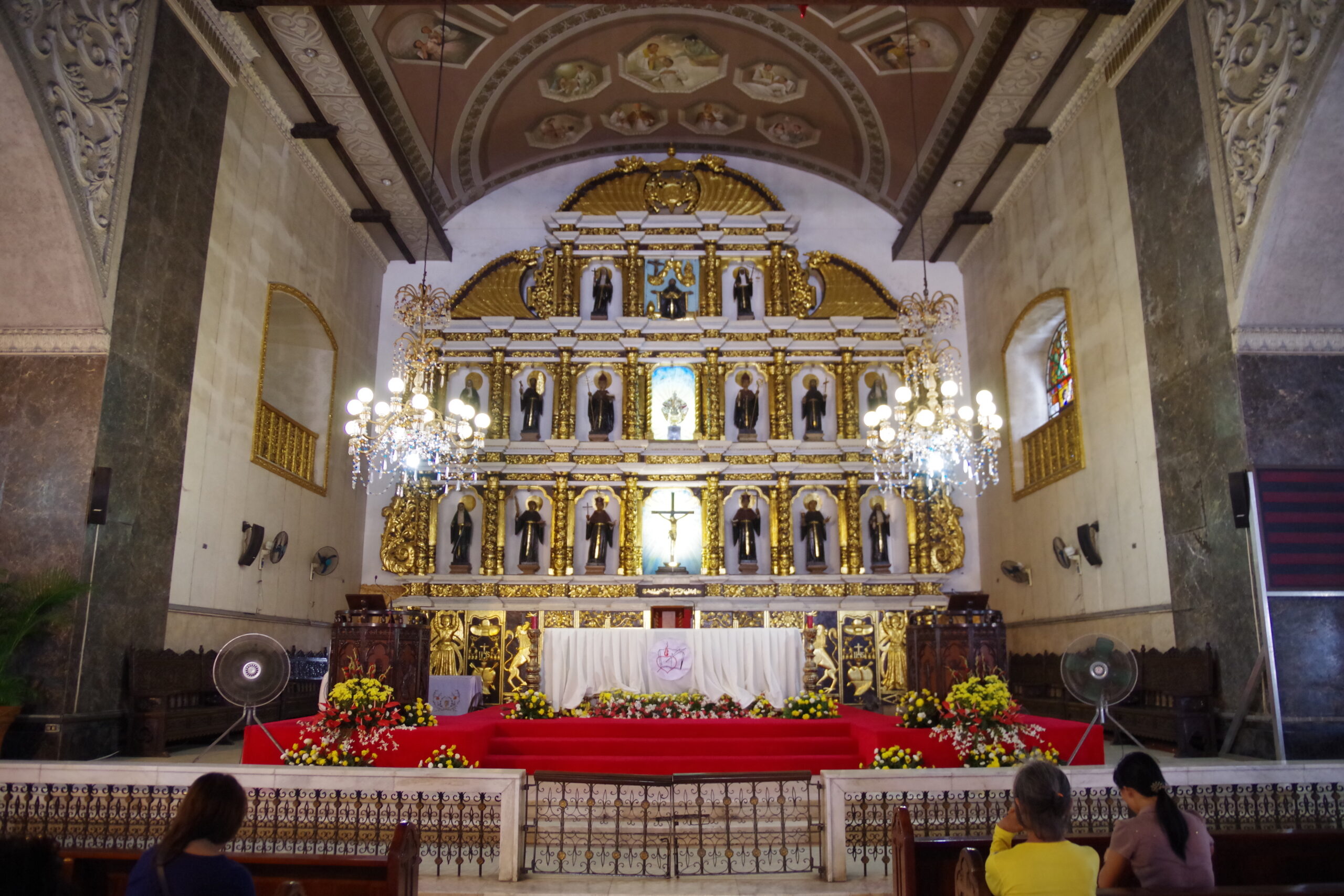 Interior of a church with a gilded, multi-tiered altar adorned with statues and icons, red carpeted steps, and chandeliers.