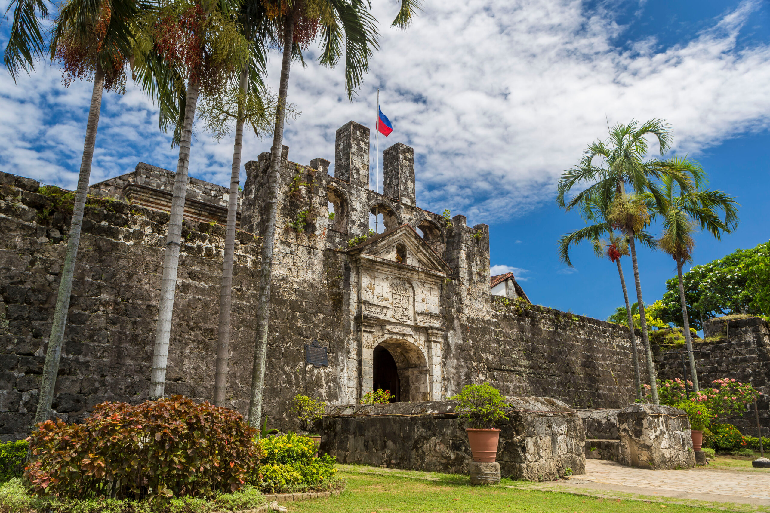 Historic stone fortress with an arched entrance, palm trees, and a flag atop a central tower against a blue sky.