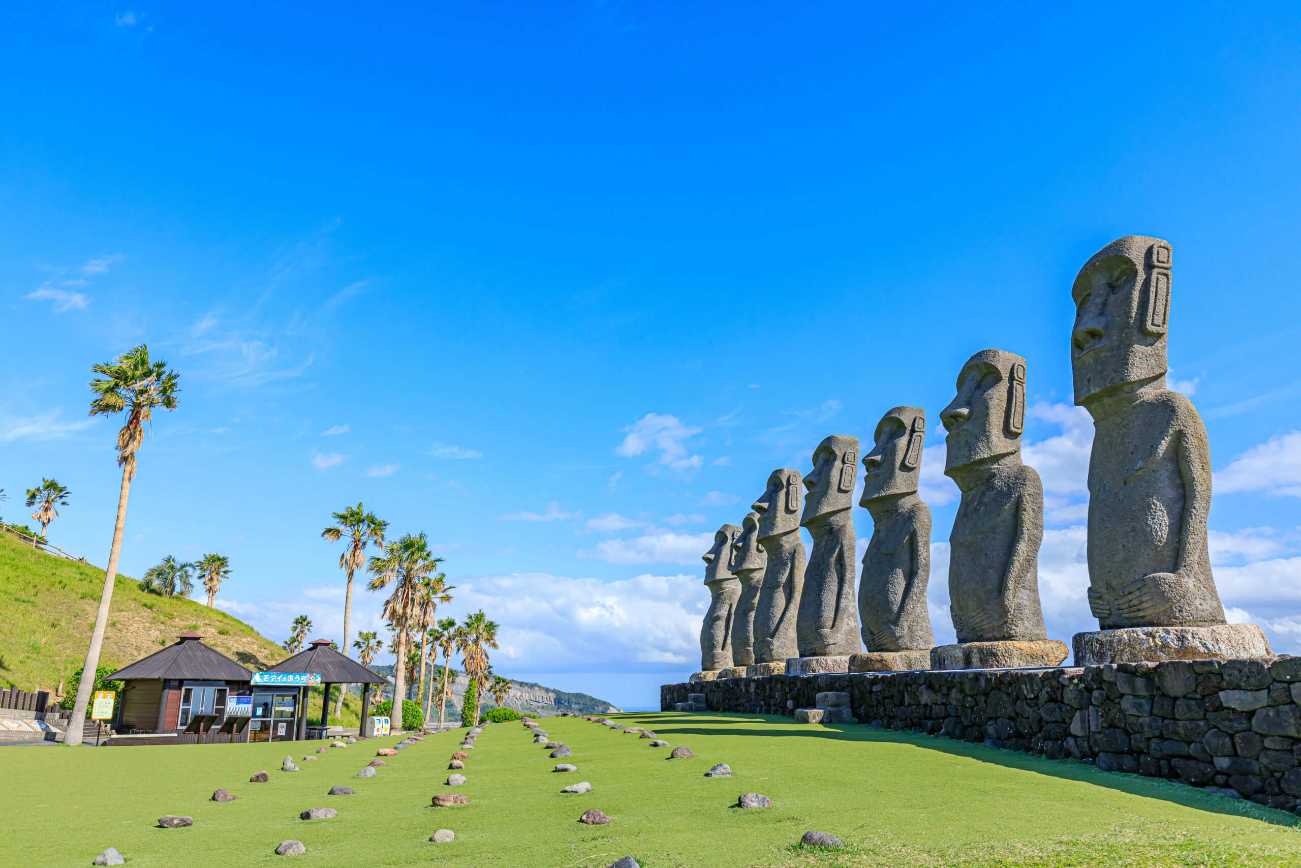 Row of large Moai statues on a stone platform under a clear blue sky, with palm trees and a small building nearby.