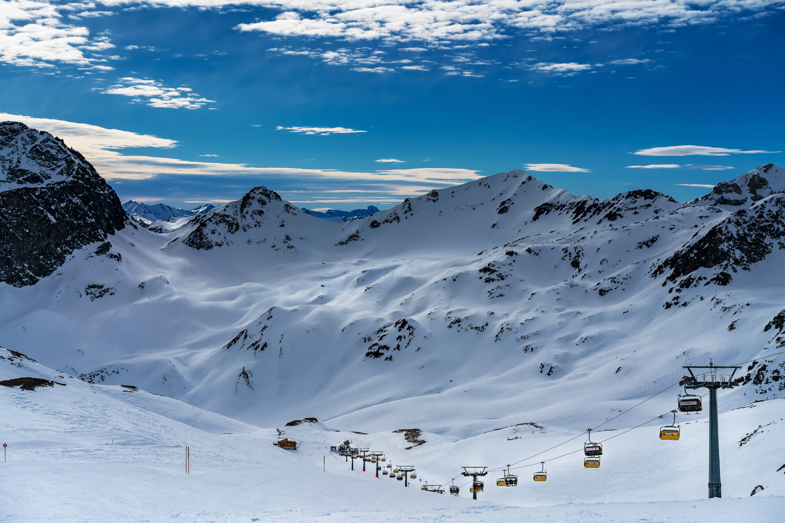 Snowy alpine valley with a chairlift running up the slope under a blue sky with clouds outskirts in the distance.