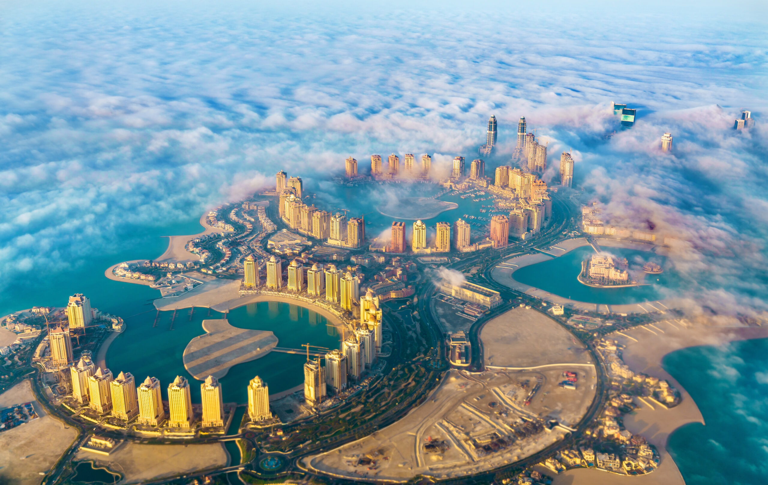 Aerial view of a modern coastal city with tall skyscrapers emerging above a ring-shaped road and blue water, surrounded by clouds.