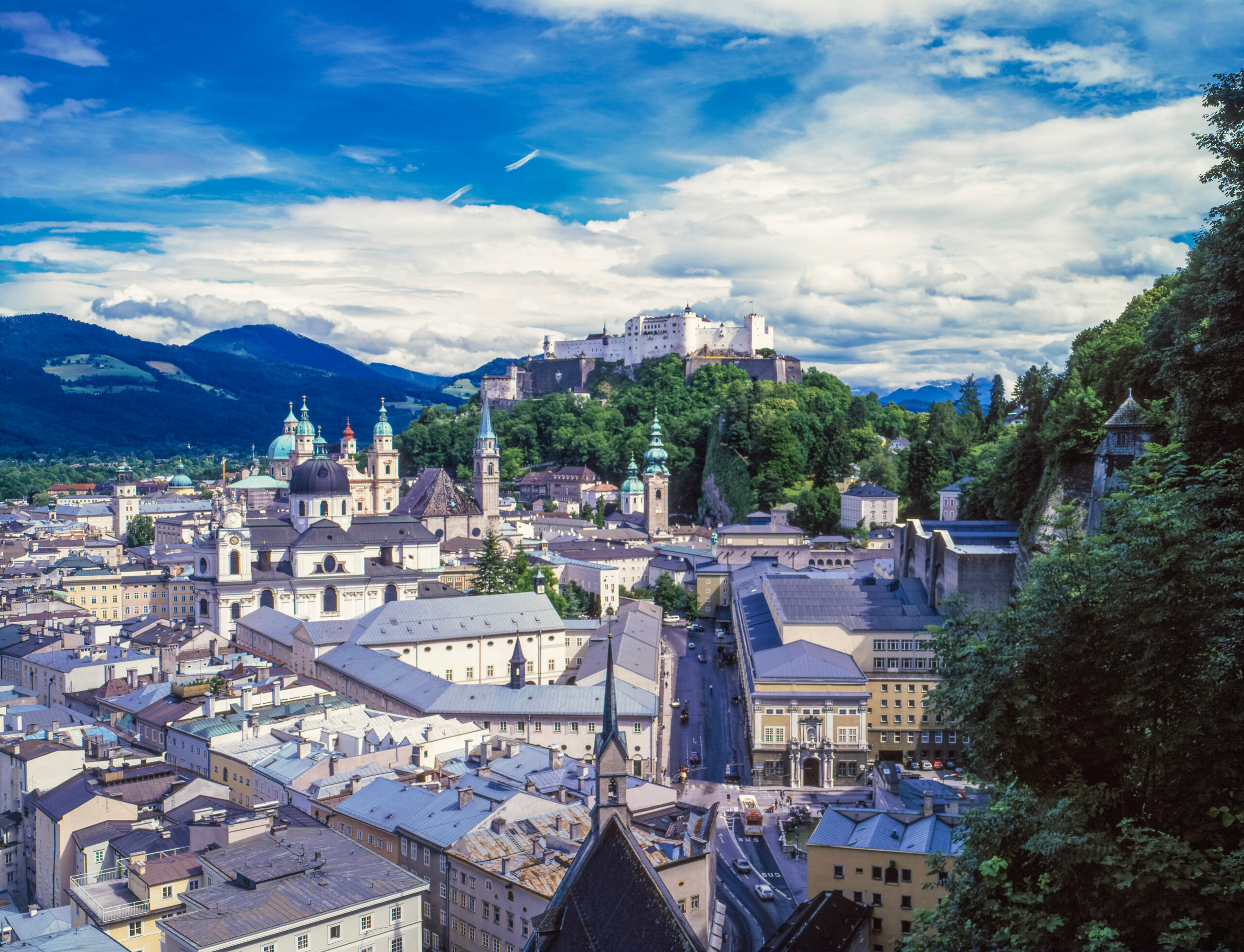 Panoramic view of a historic European city with a hilltop fortress, church domes, and dense rooftops under a bright blue sky above distant hills.